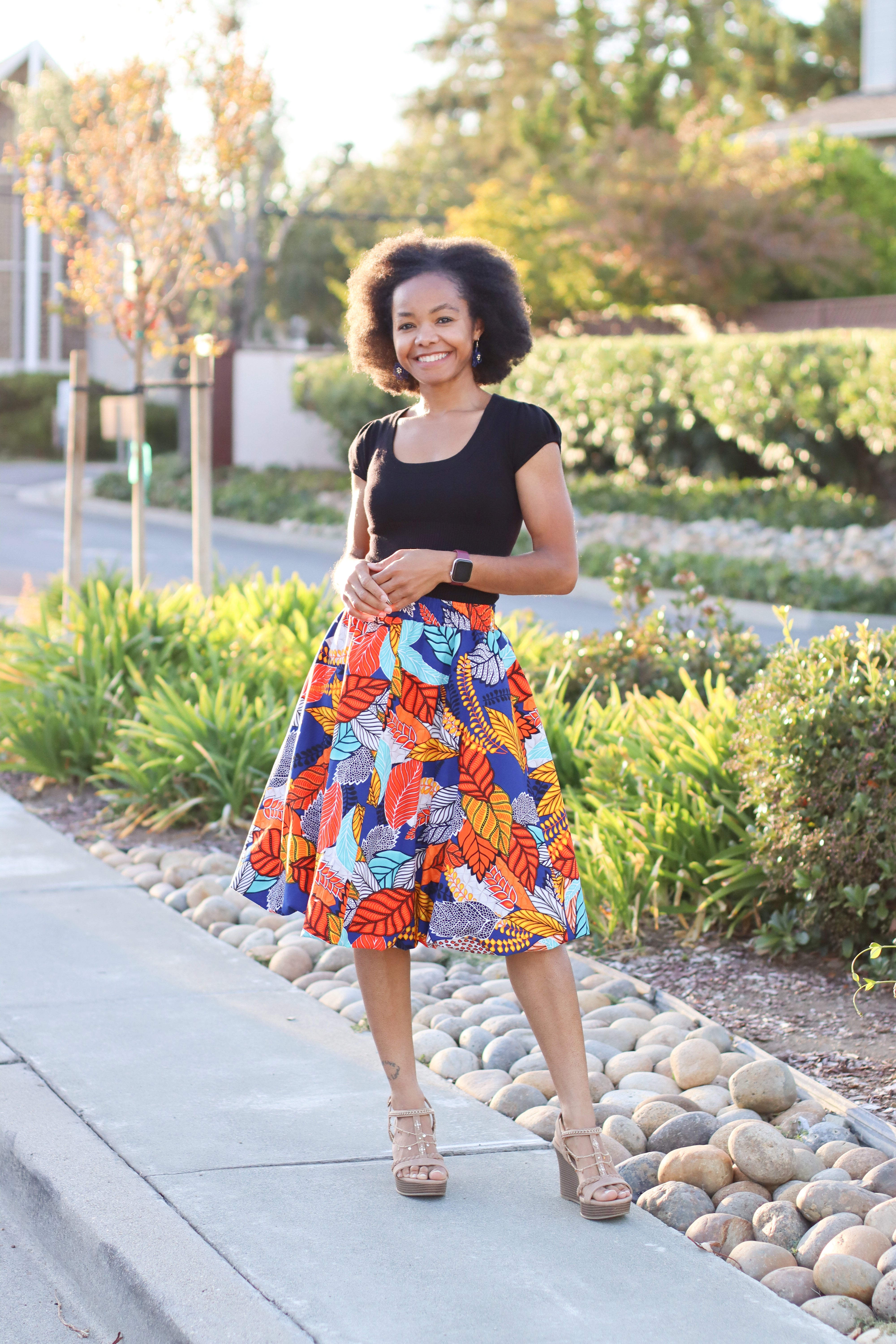Woman in a colorful knee-length bubble skirt standing on a sidewalk with greenery in the background. Skirt is made using Ellie and Mac's Sadie Bubble Skirt PDF sewing pattern.