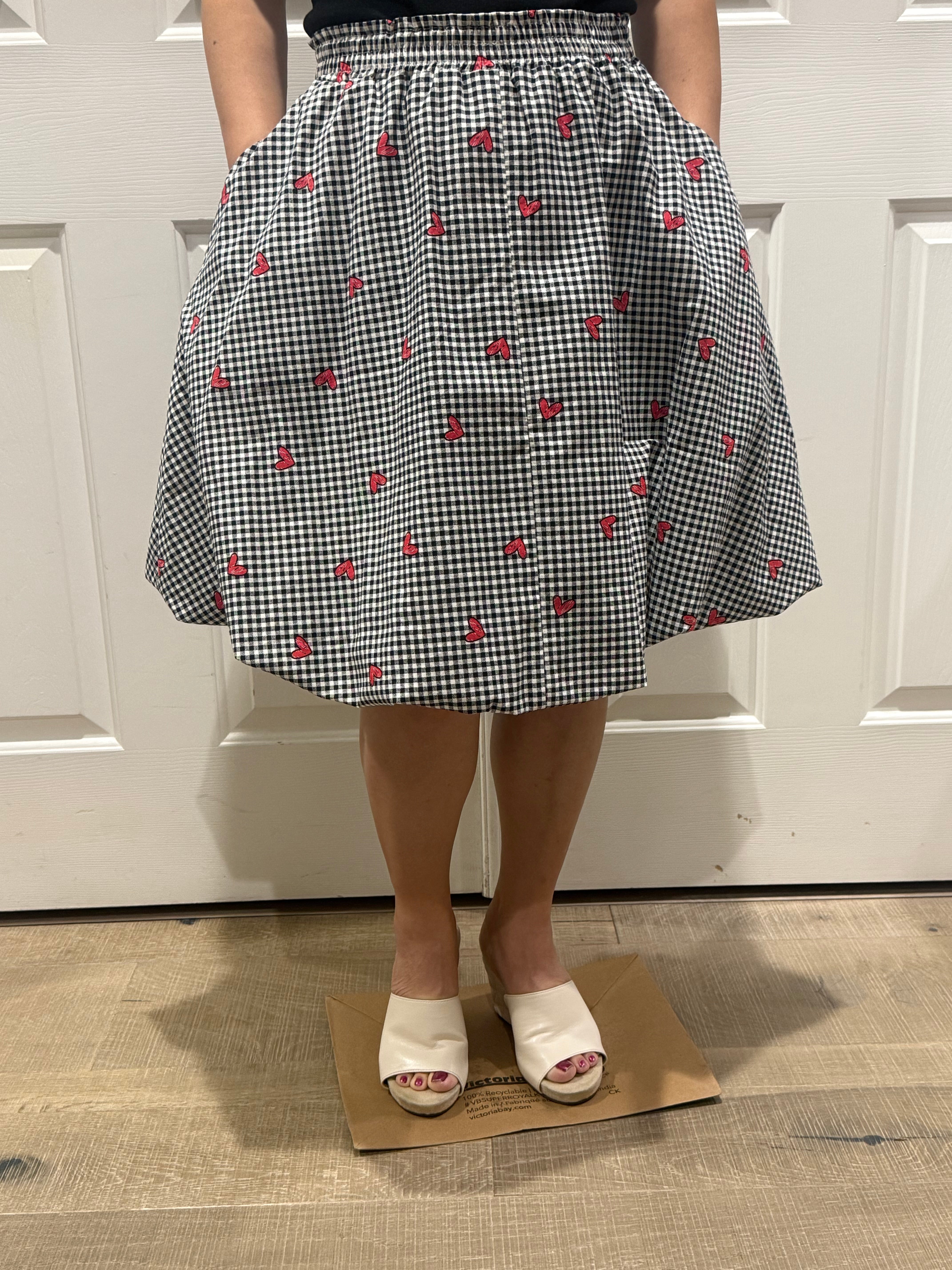 Person wearing a black and white checkered apron with red patterns, standing on a wooden floor. Make it with the Sadie Bubble Skirt pattern.