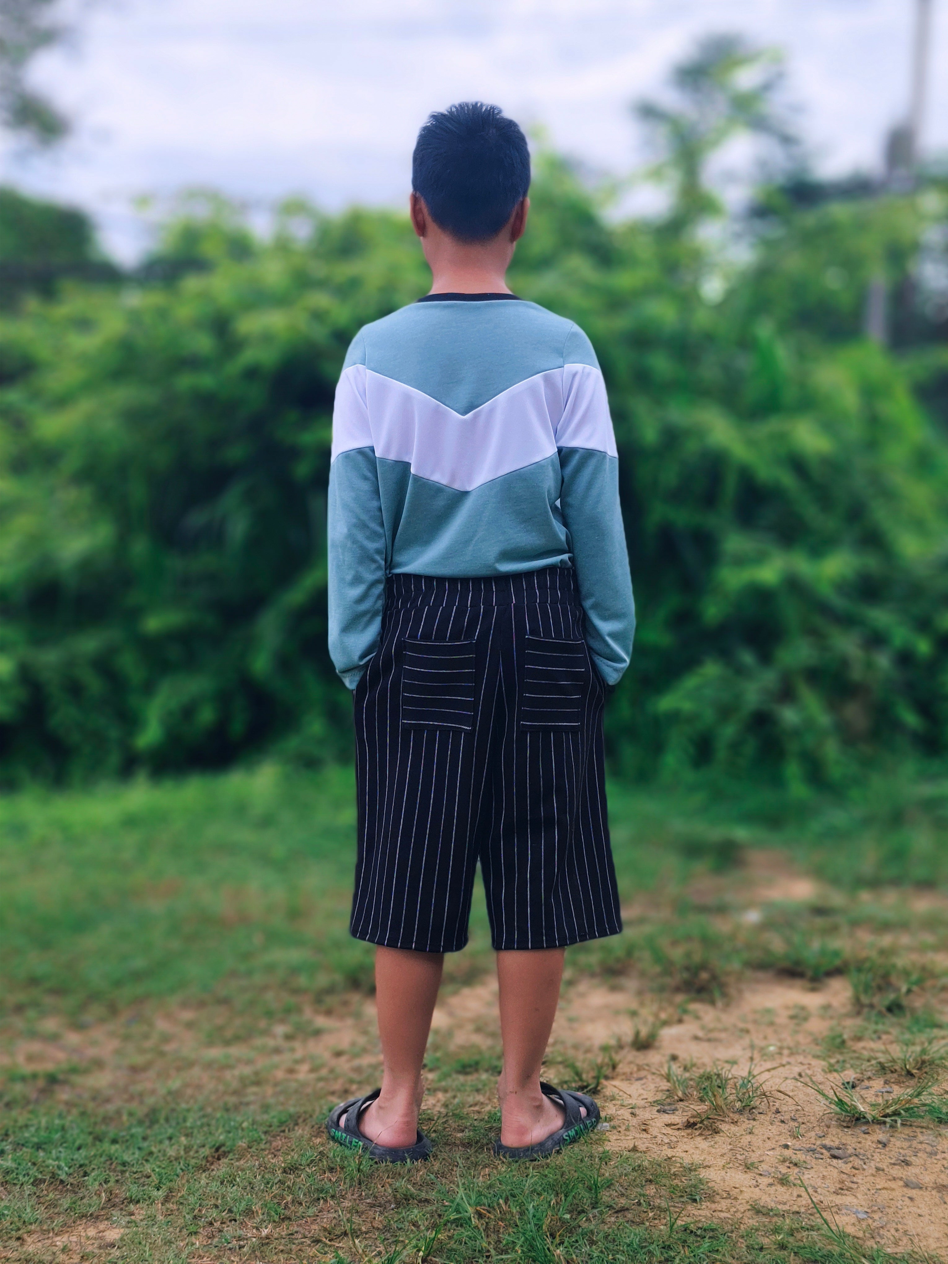 Boy standing in an outdoor setting wearing a teal and white color block shirt and striped knee length shorts made using the Caleb Athletic Sewing Pattern by Ellie and Mac Patterns.