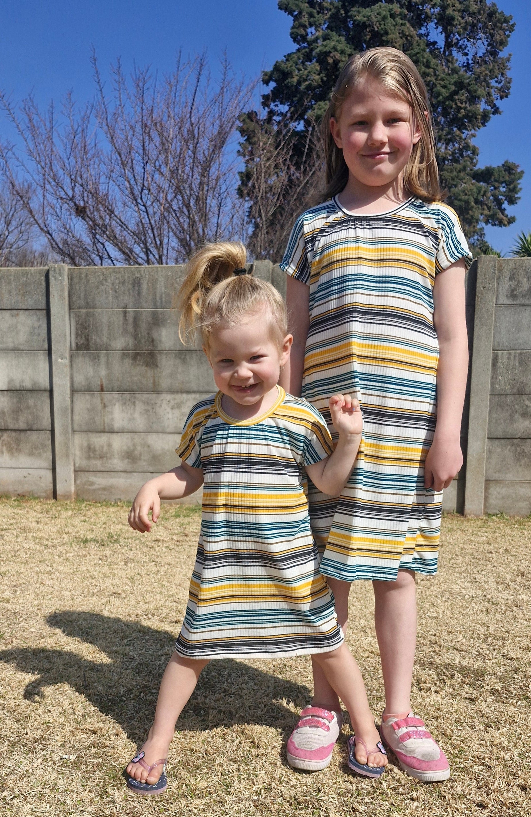 Two children in matching striped dresses standing on a gravel path with a clear blue sky. Dresses made using the Kayla Crop Top and Dress sewing pattern by Ellie and Mac patterns.