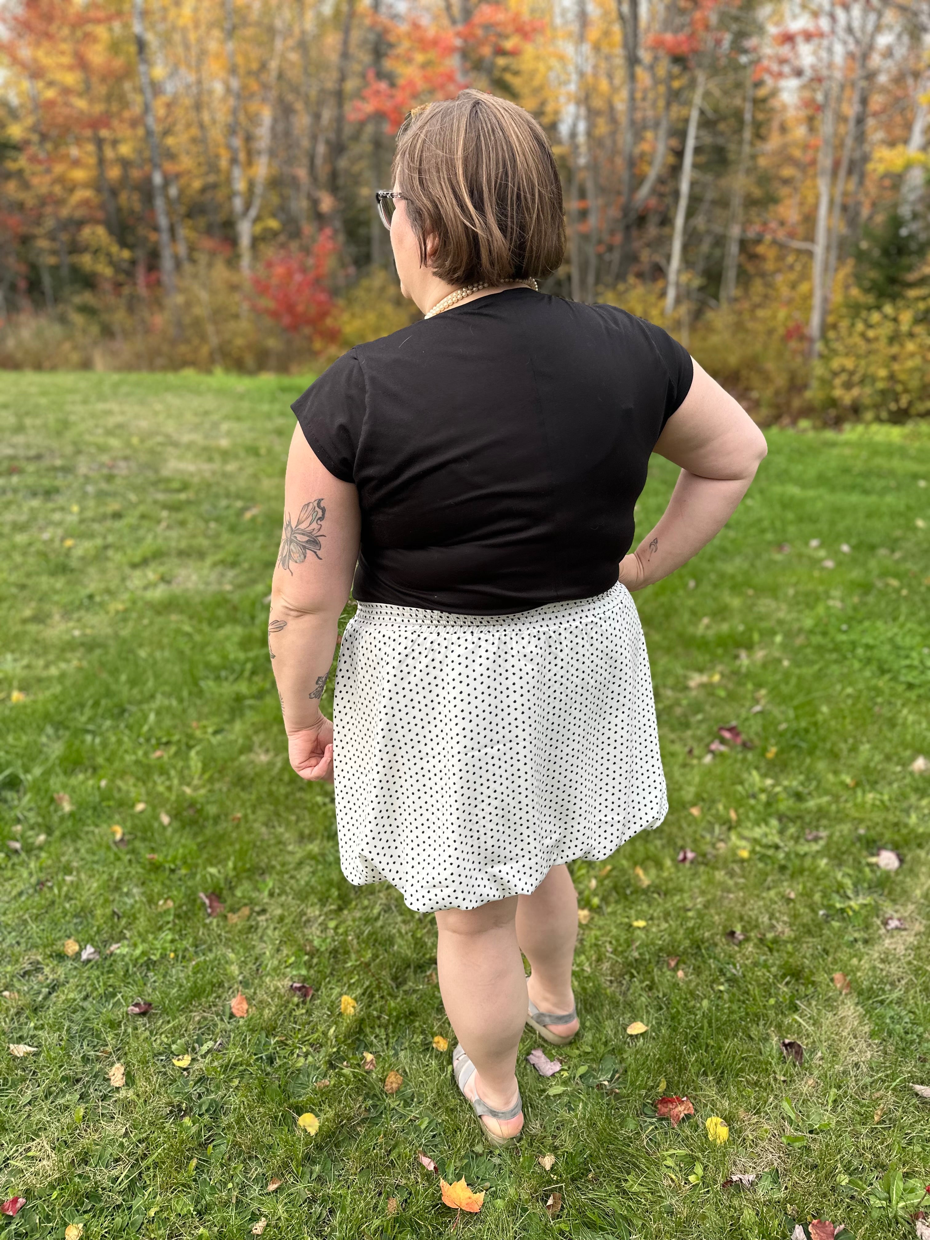 Person wearing a black t-shirt and white polka dot bubble skirt standing in a grassy area with trees in the background.
