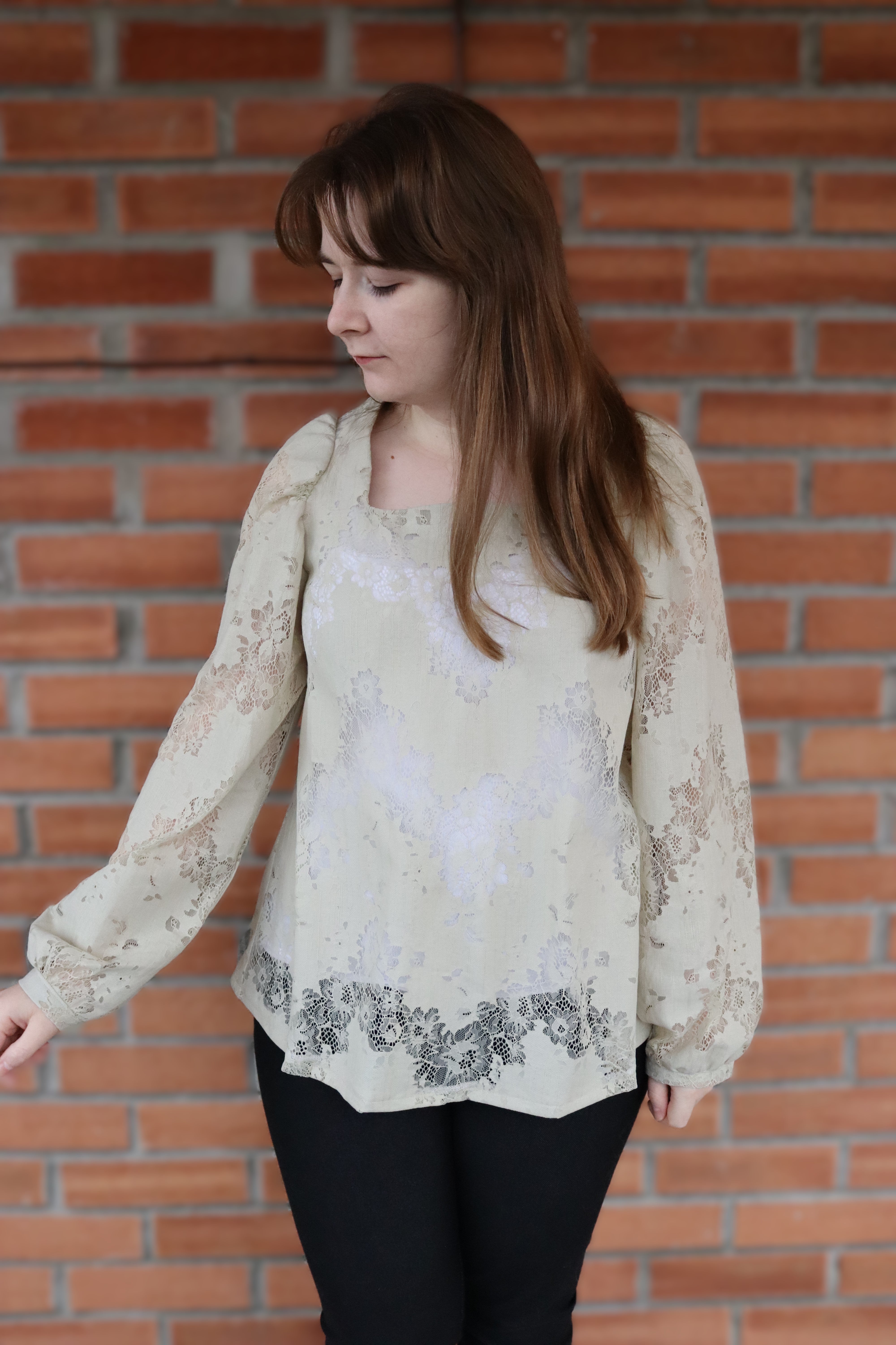Woman wearing a beige lace Zara Square Neck Blouse against a brick wall with black pants.