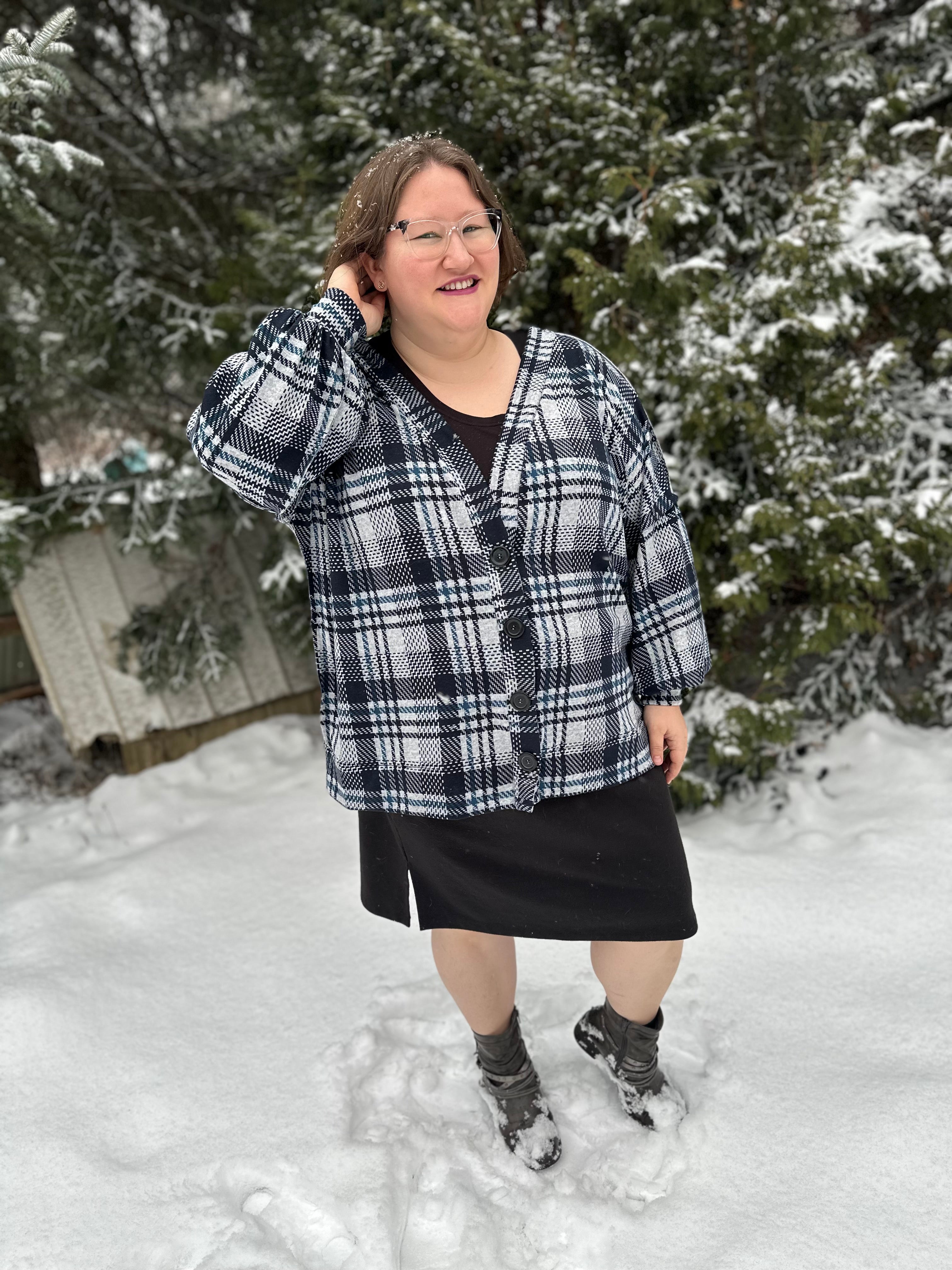 Person wearing a plaid oversized cardigan and black skirt standing in the snow with trees in the background.