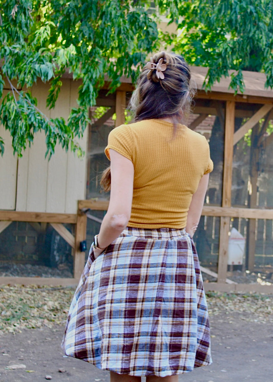 Person wearing a mustard yellow top and plaid skirt she made using the Sadie Bubble Skirt Pattern standing outdoors with greenery in the background.