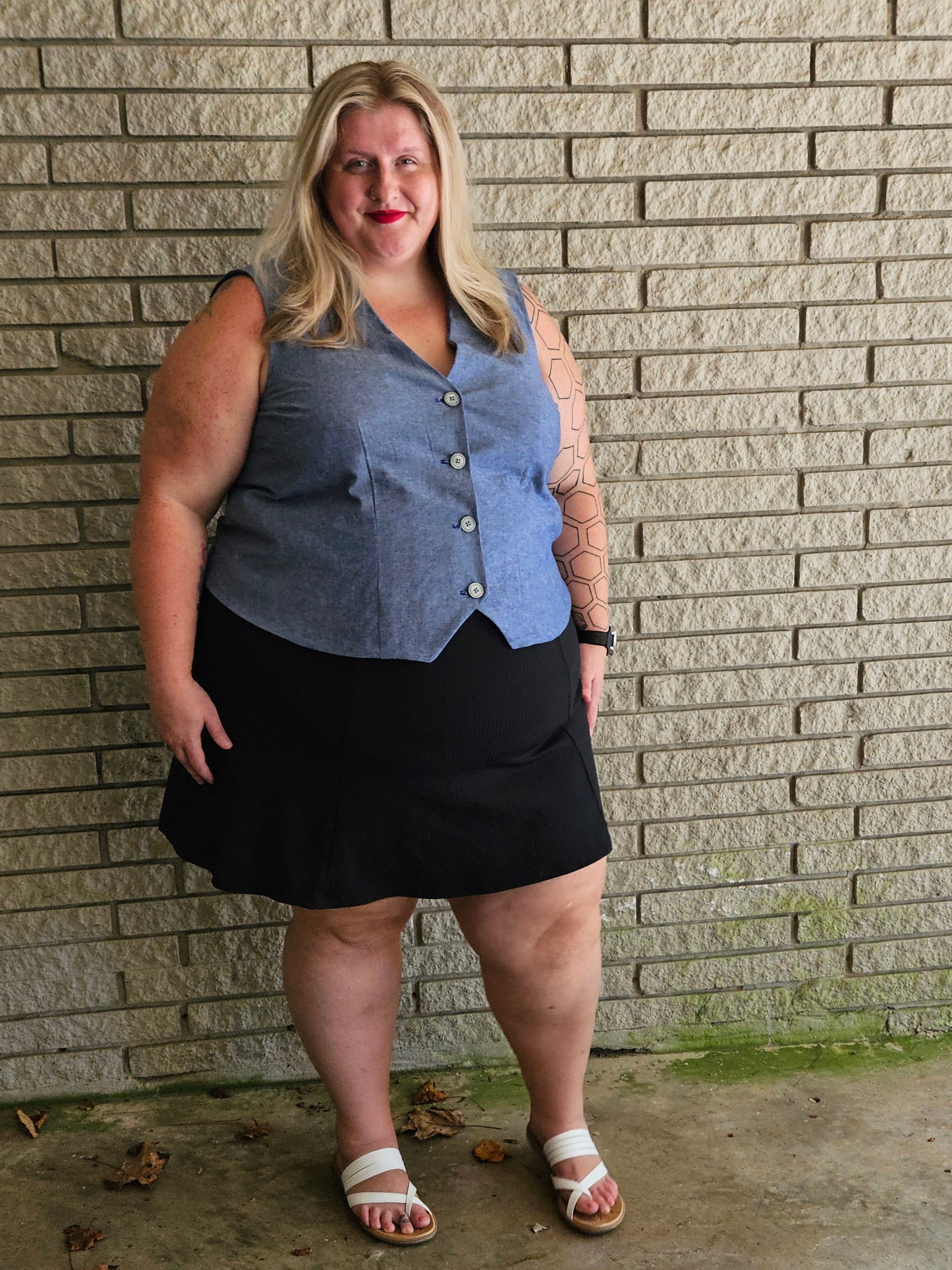 Woman wearing a blue waistcoat and black skirt standing against a brick wall.