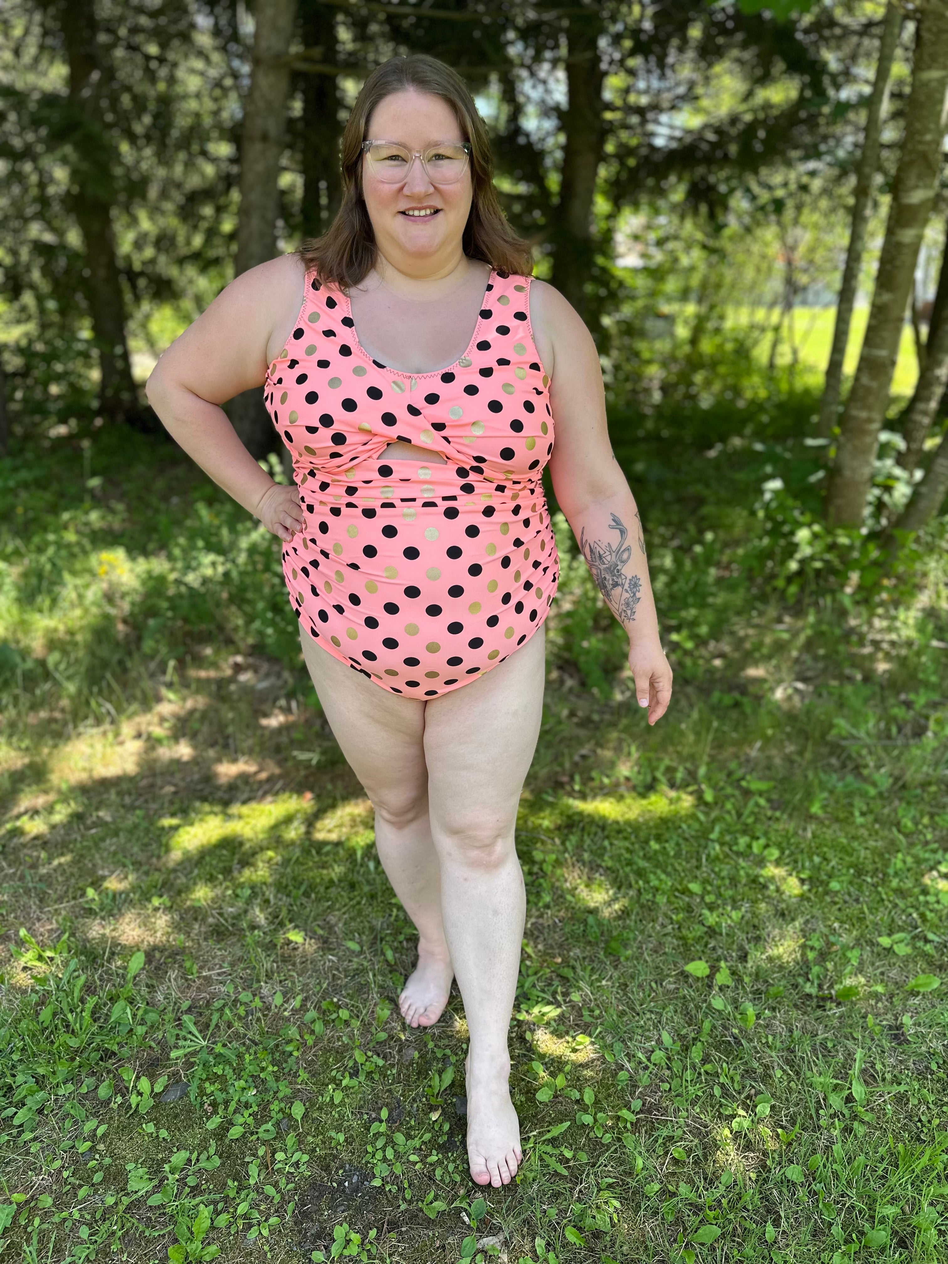 Person wearing a pink polka dot swimsuit standing in a grassy area with trees.