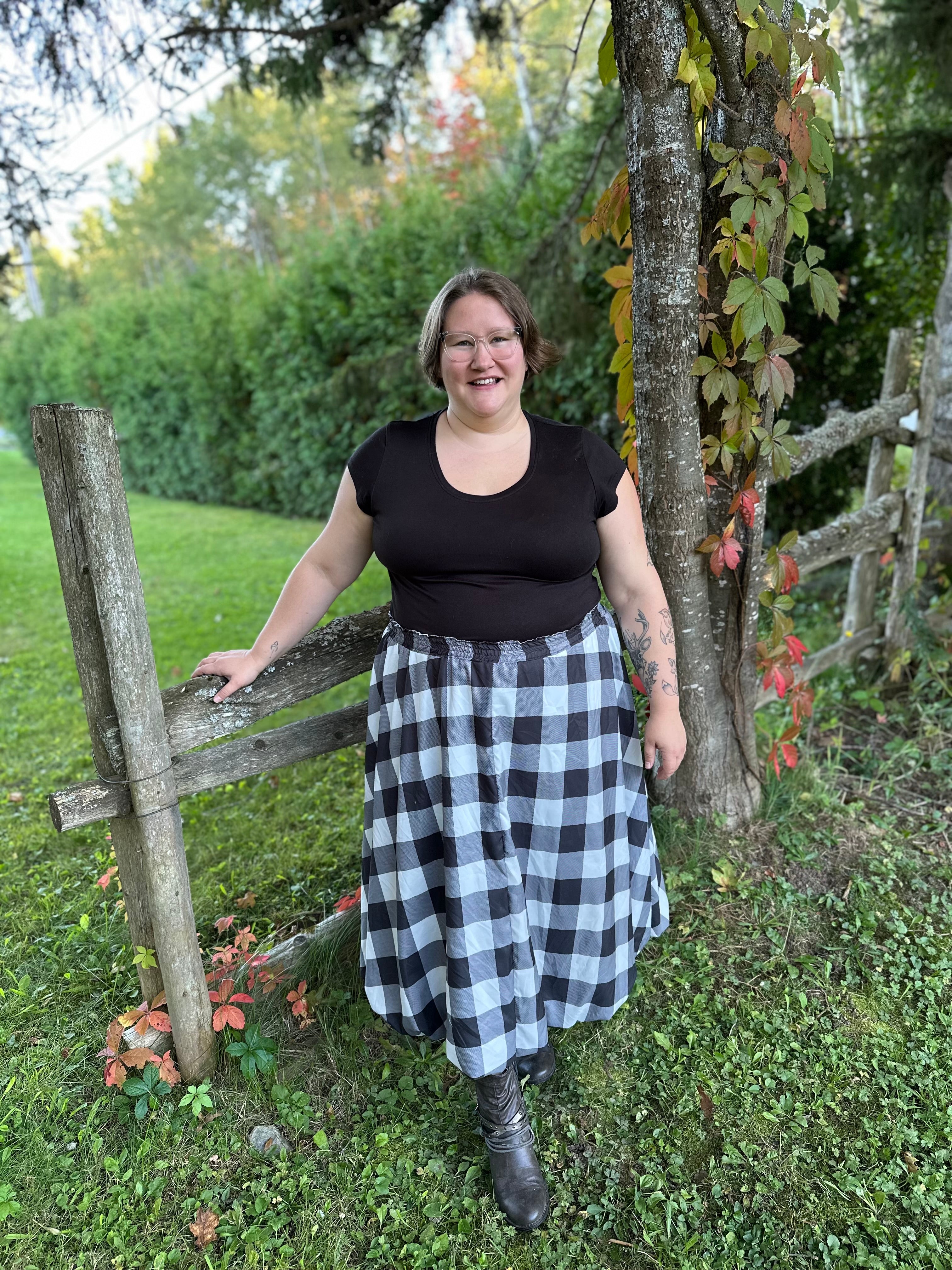 Woman wearing a handmade buffalo plaid ankle bubble skirt, standing outdoors by a wooden fence with trees, and grass in the background.