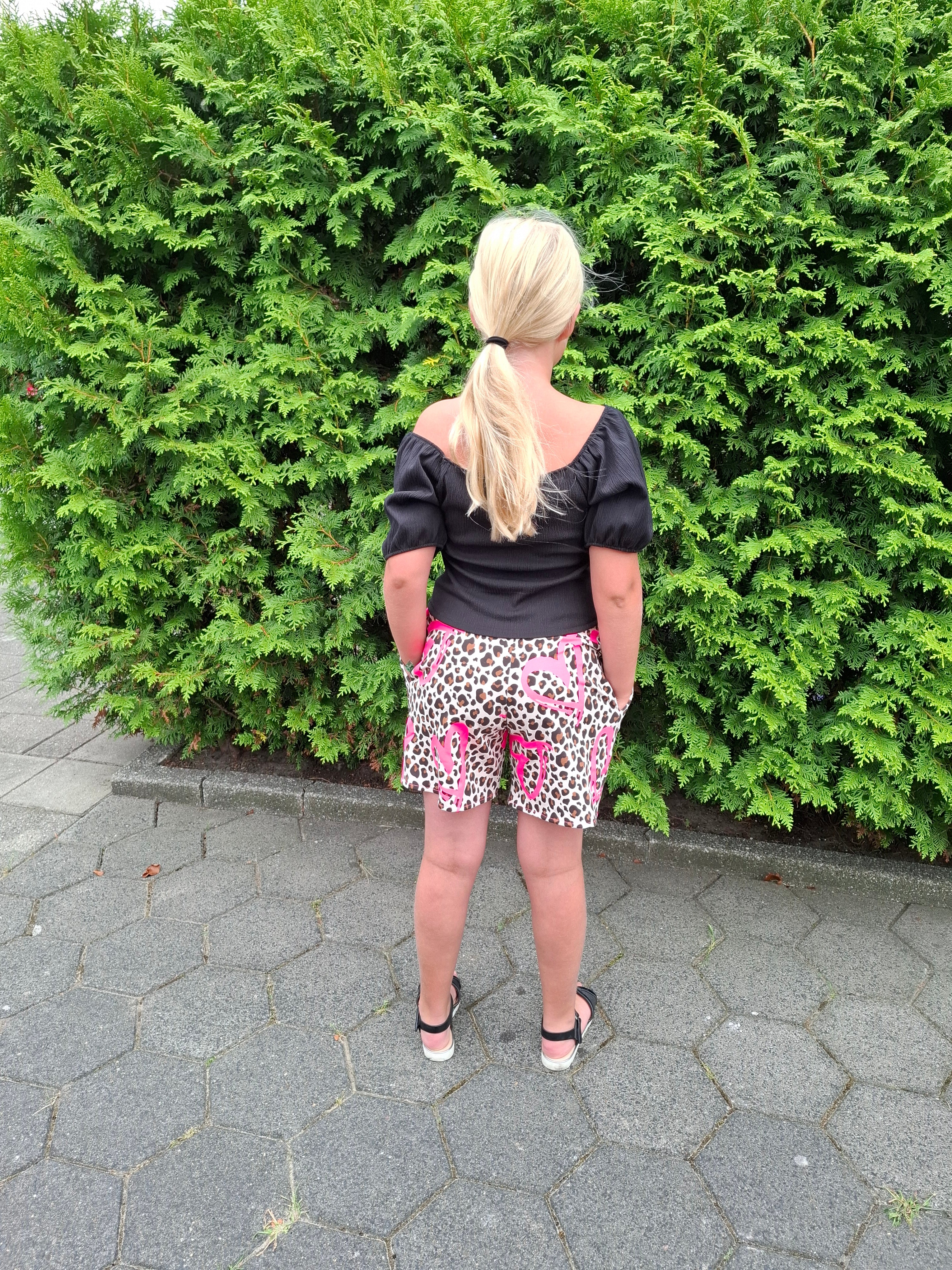 Girl standing next to a green bush on a paved path with blond hair, a black top, and cute shorts.