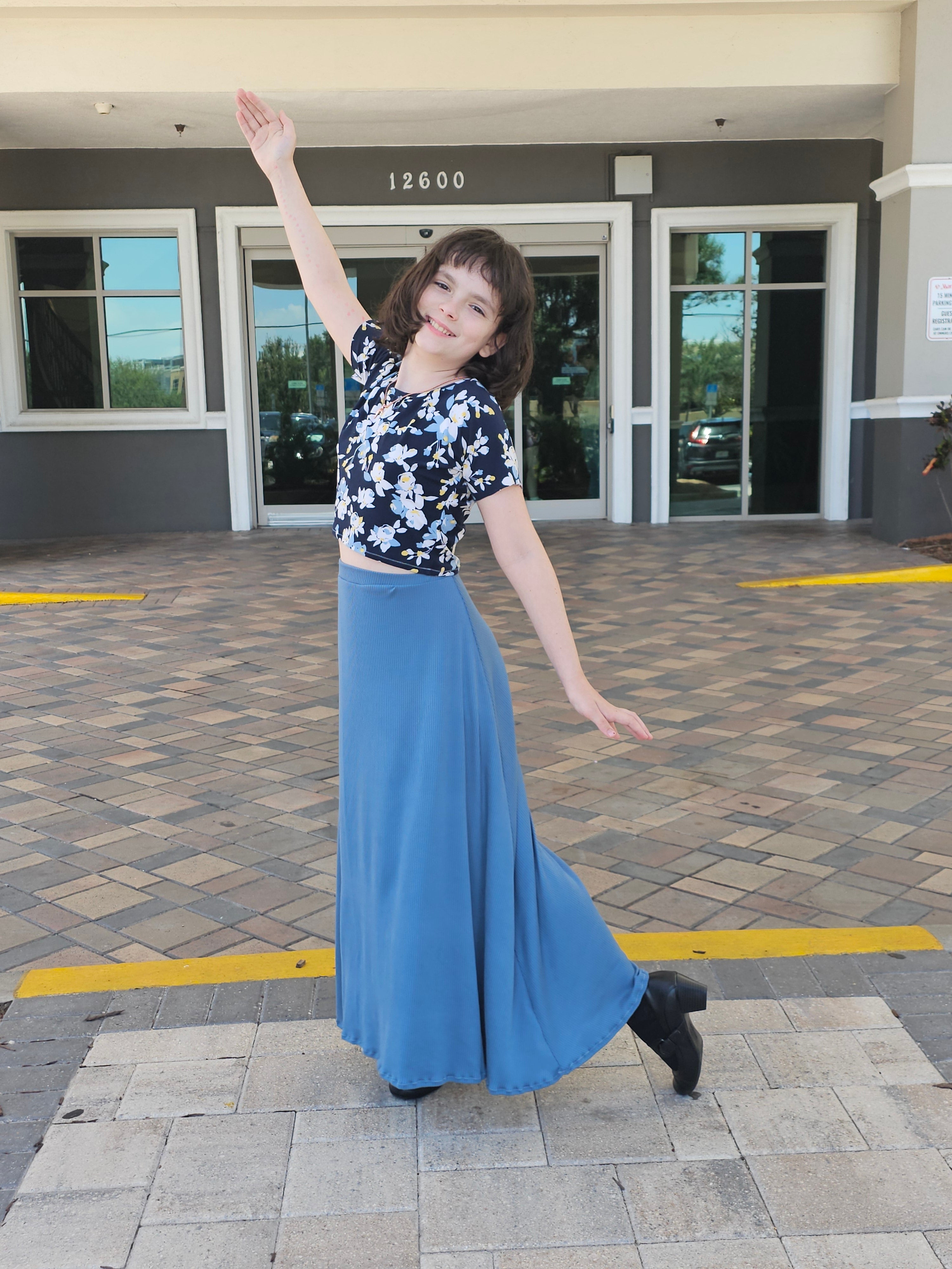 Woman in a floral top and blue skirt standing on a sidewalk with a building in the background