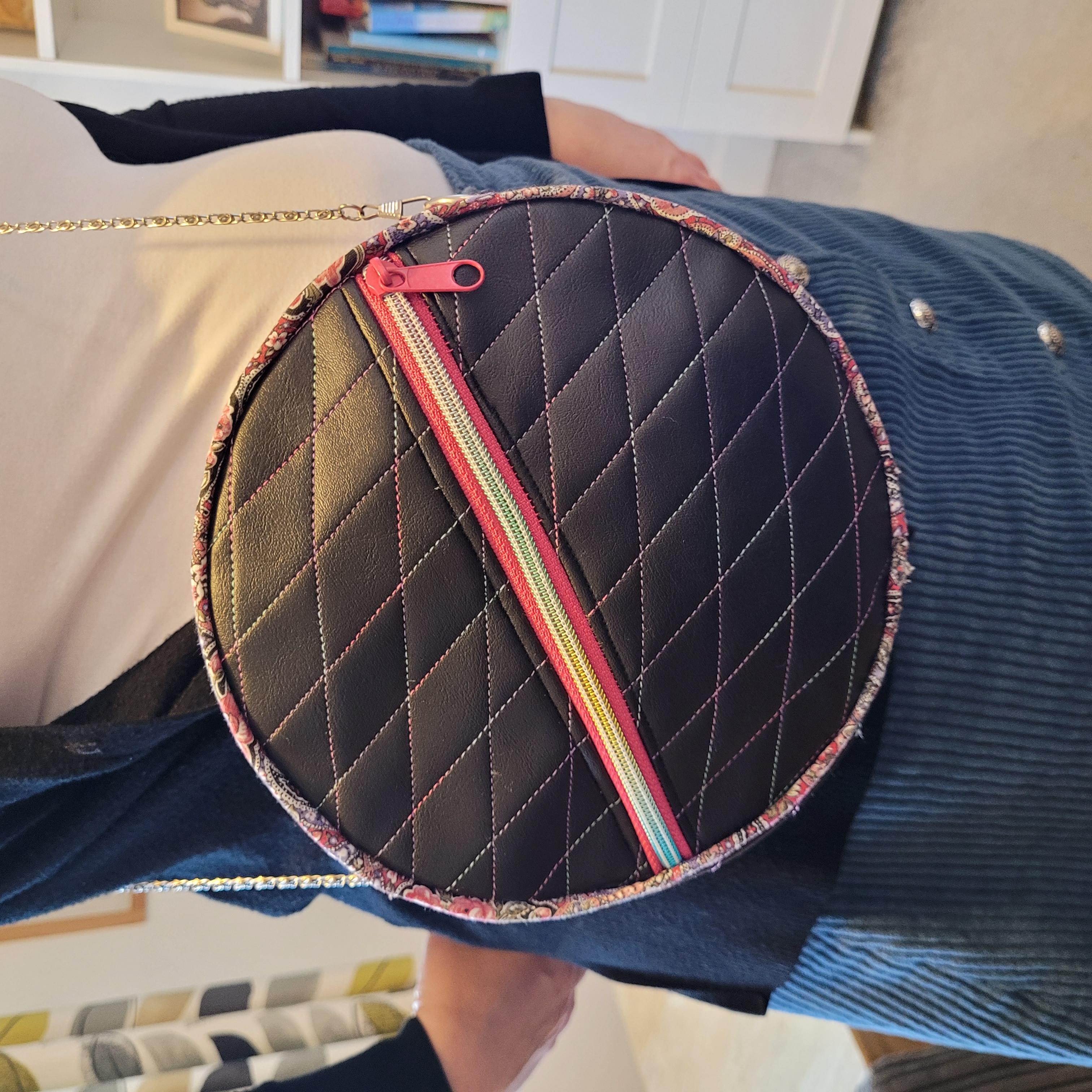 Round quilted bag with a zipper held by a person in a casual setting.