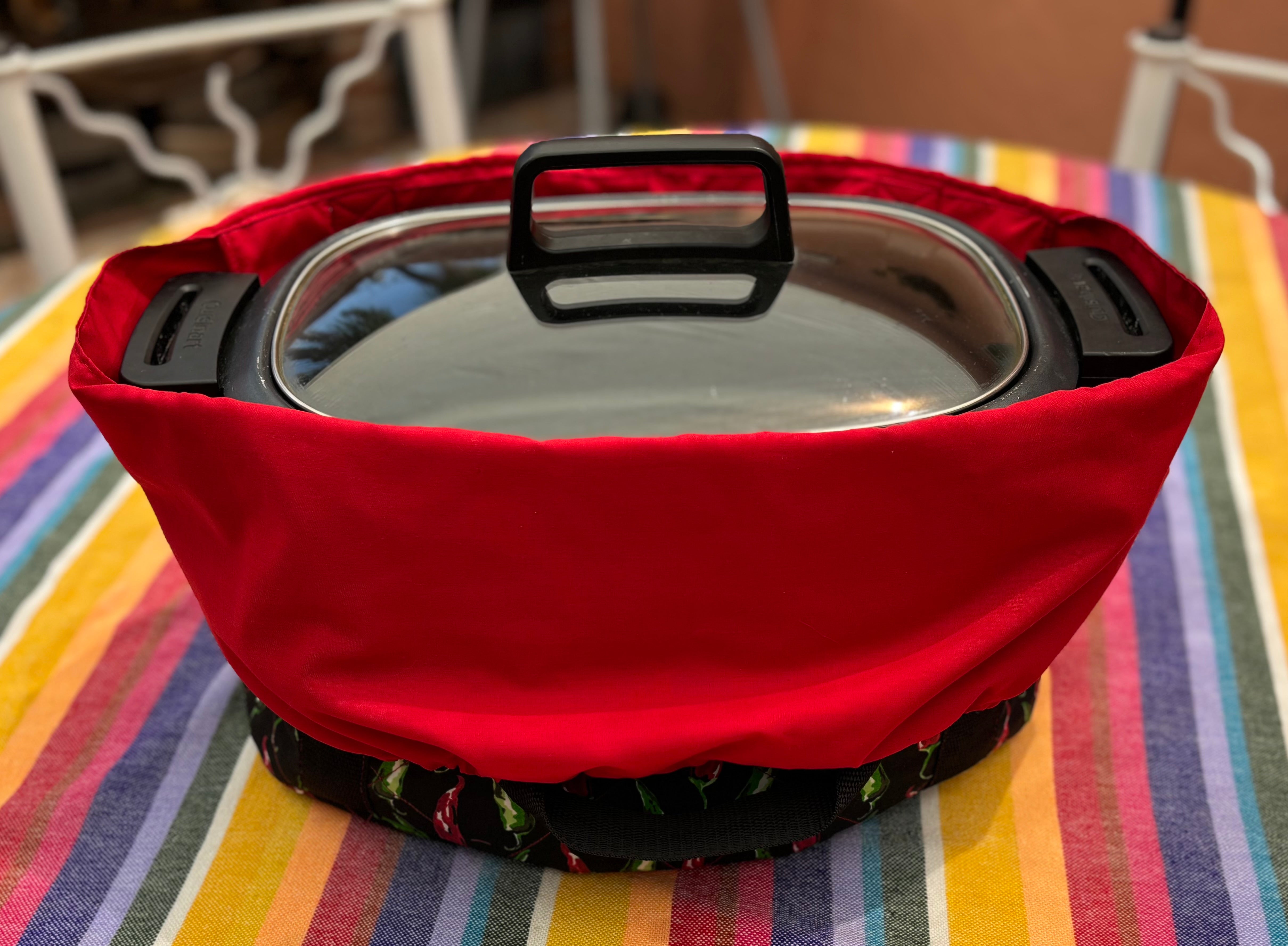 Red crock pot holder with a pot on a colorful tablecloth.