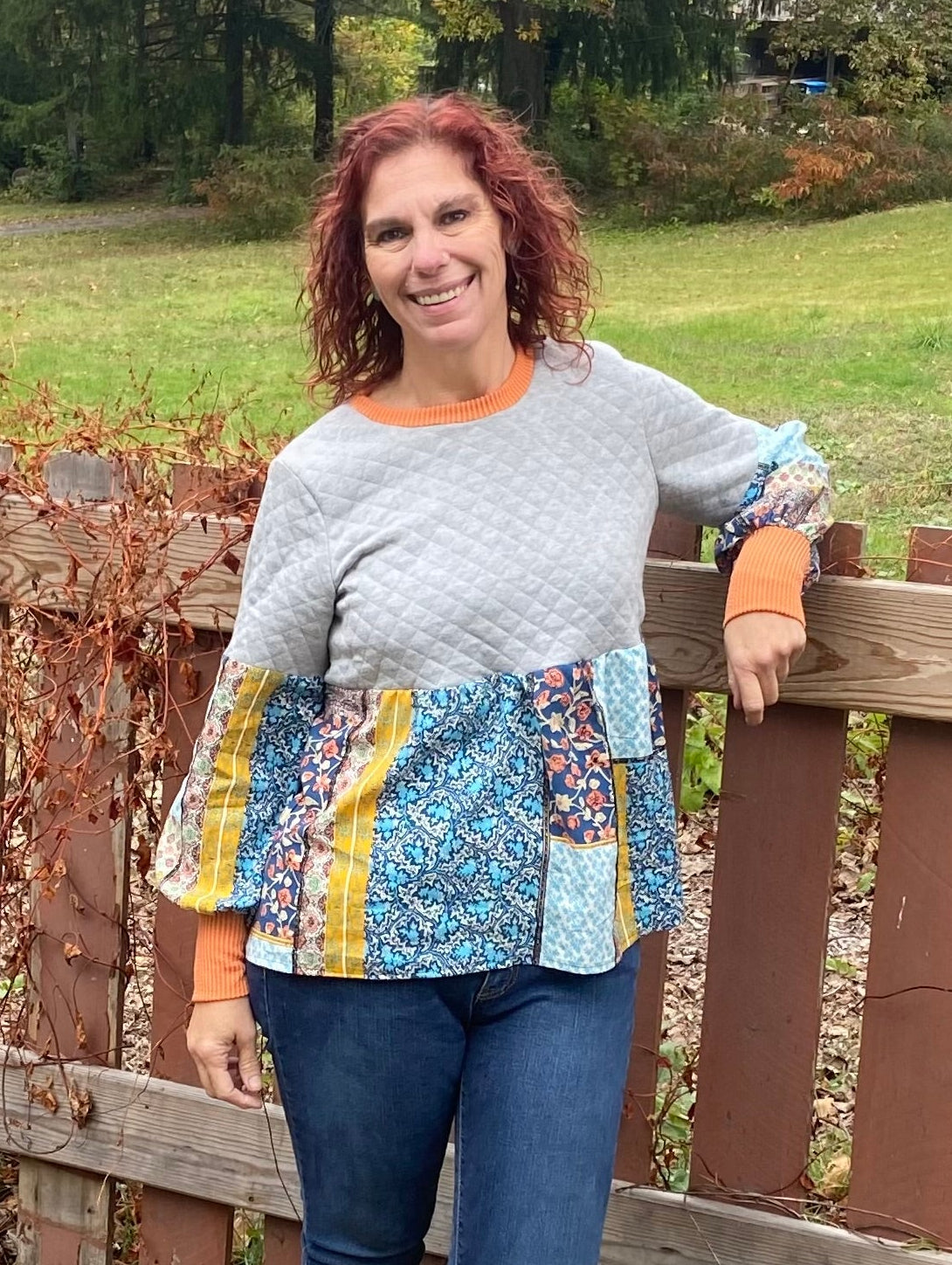 Woman standing outdoors by a wooden fence with a colorful top and blue jeans. Made using the Imogen Mixed Media Peasant Top by Ellie and Mac PDF sewing patterns.