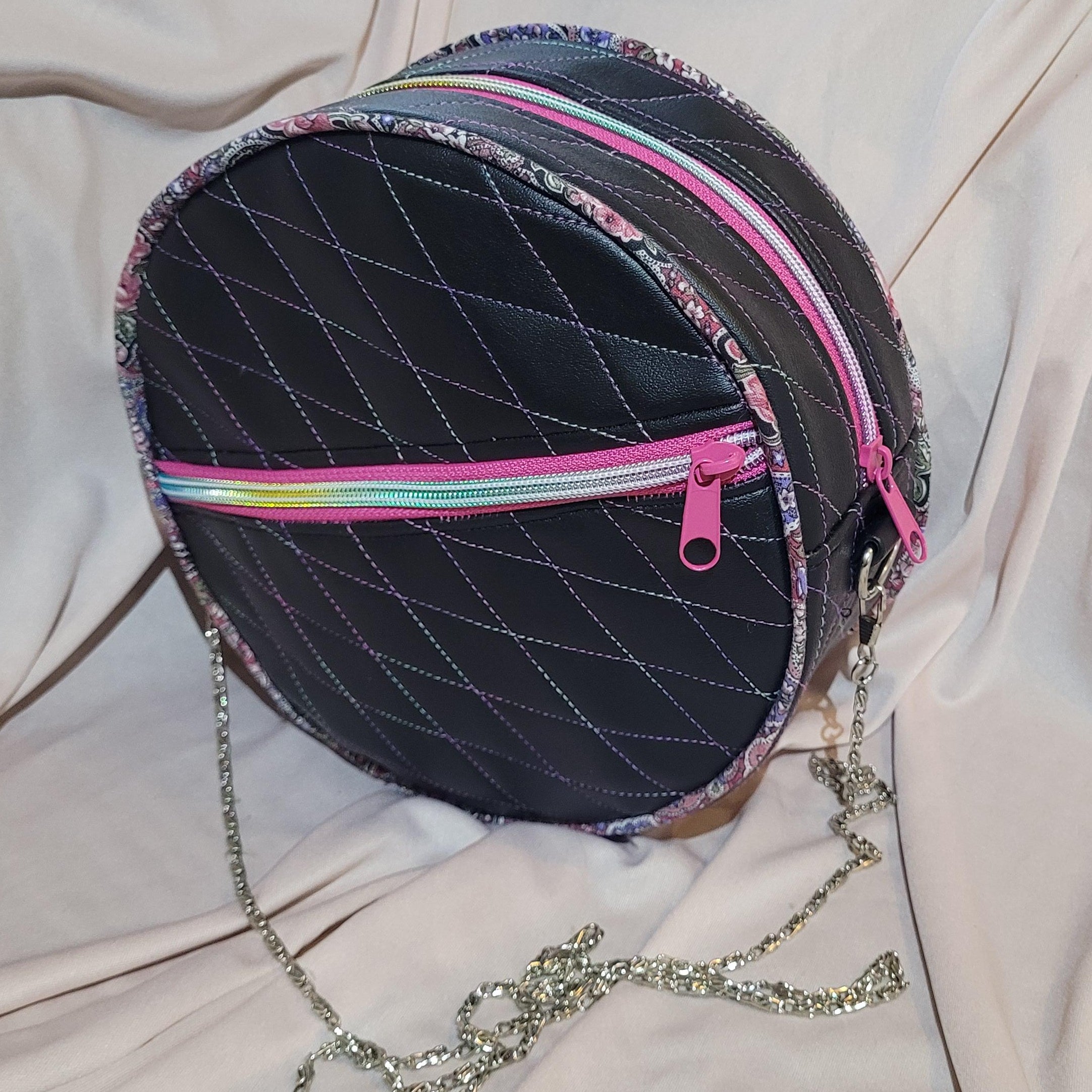 Round black bag with colorful zippers and a chain strap on a light pink fabric background