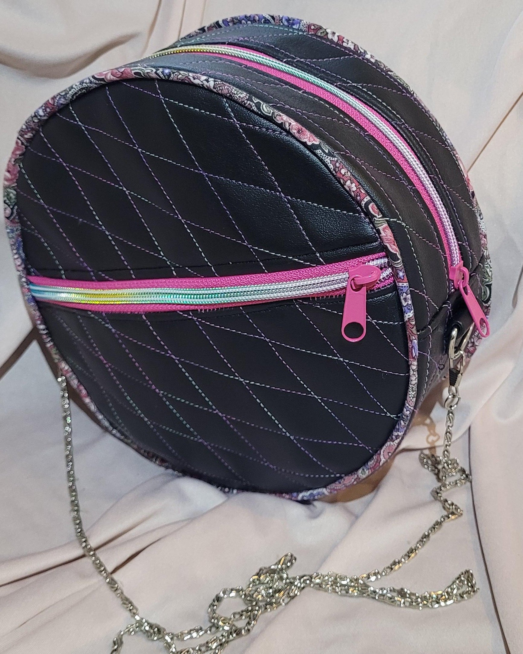Round black bag with colorful zippers and a chain strap on a light pink fabric background