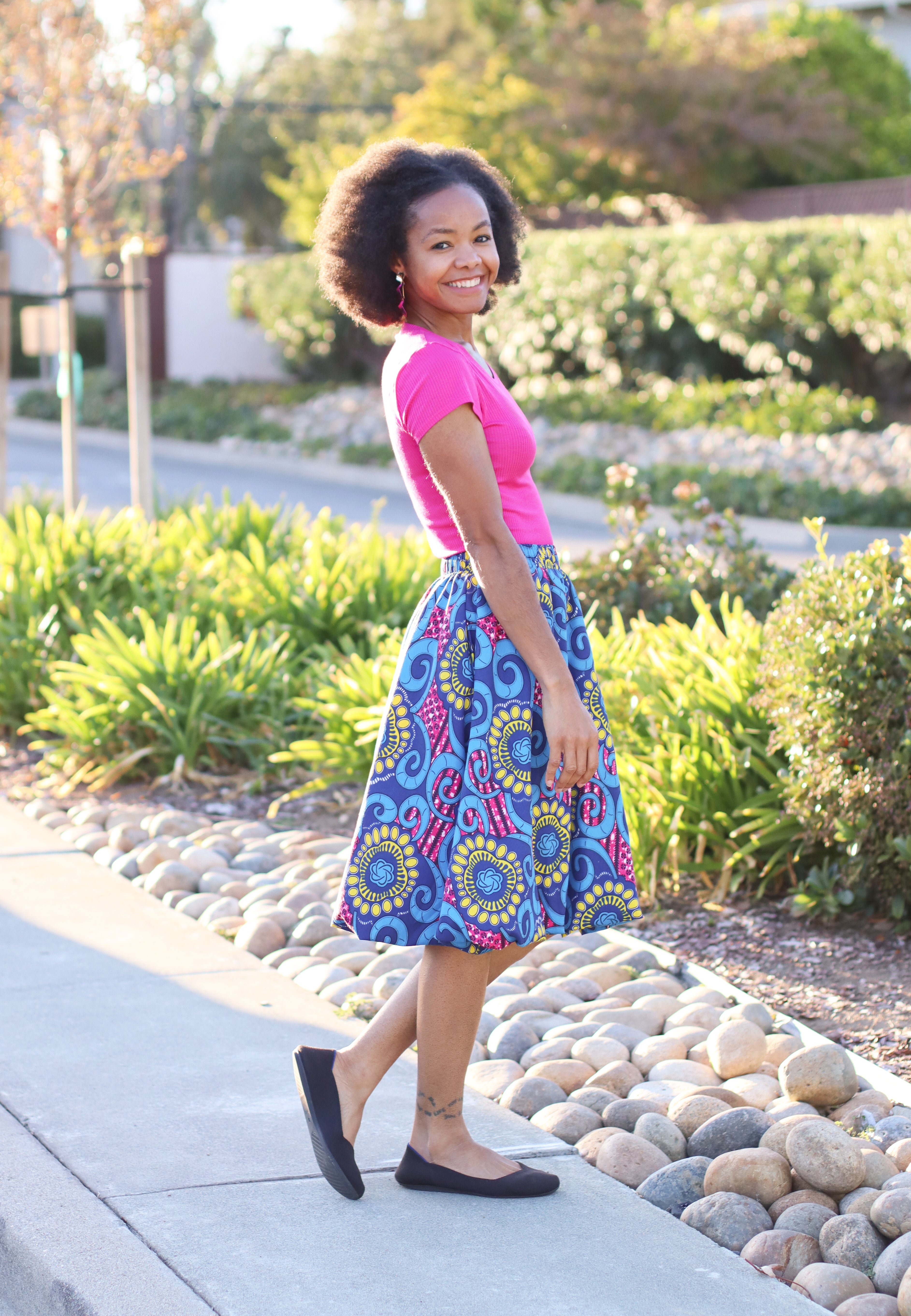 A smiling woman wearing a hot pink top and colorful bubble skirt standing outdoors on a sidewalk.