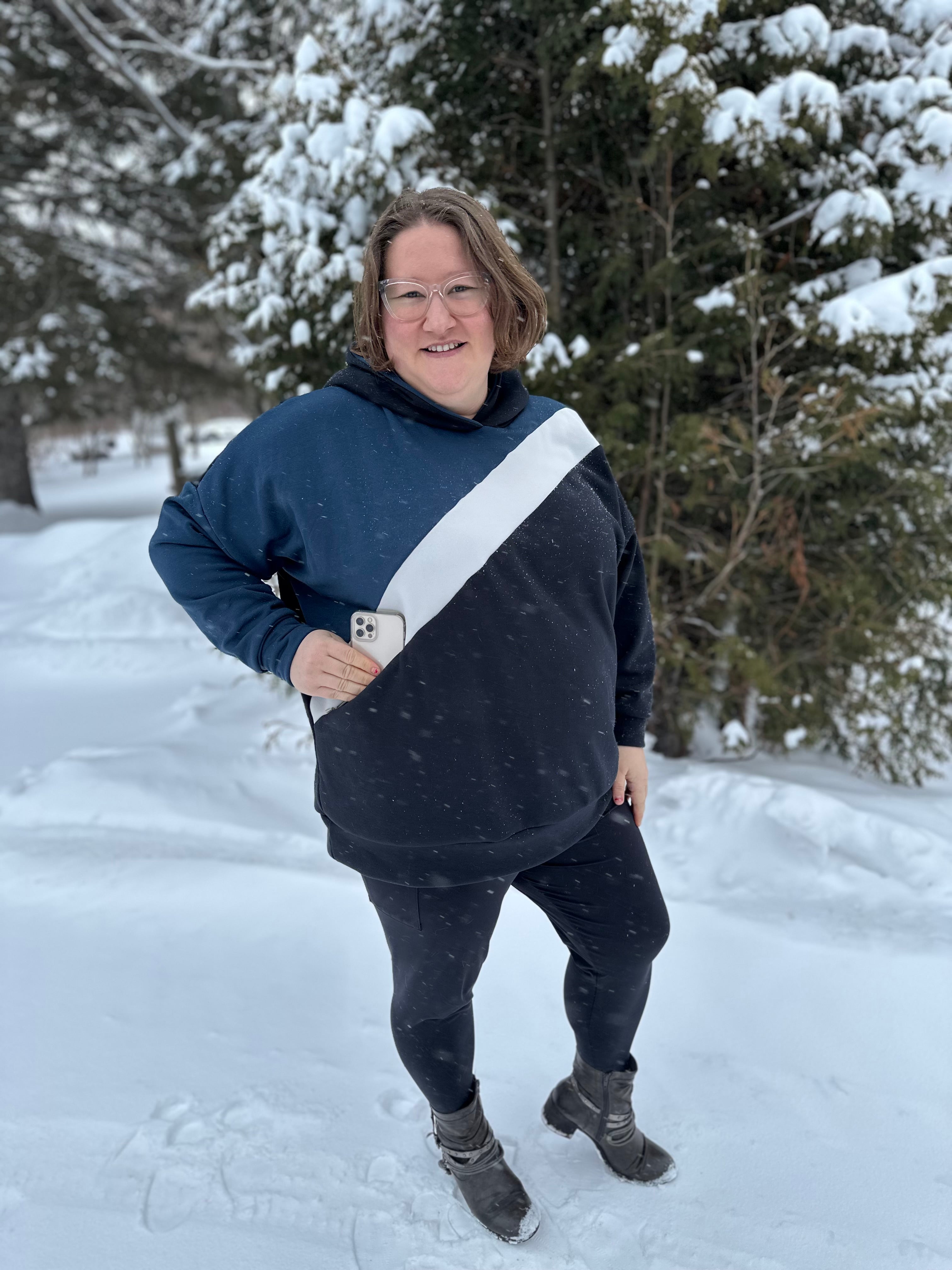 Person wearing a navy and gray hoodie with a white stripe, standing in a snowy landscape. Showing off the zip pocket with her cell phone going into it.