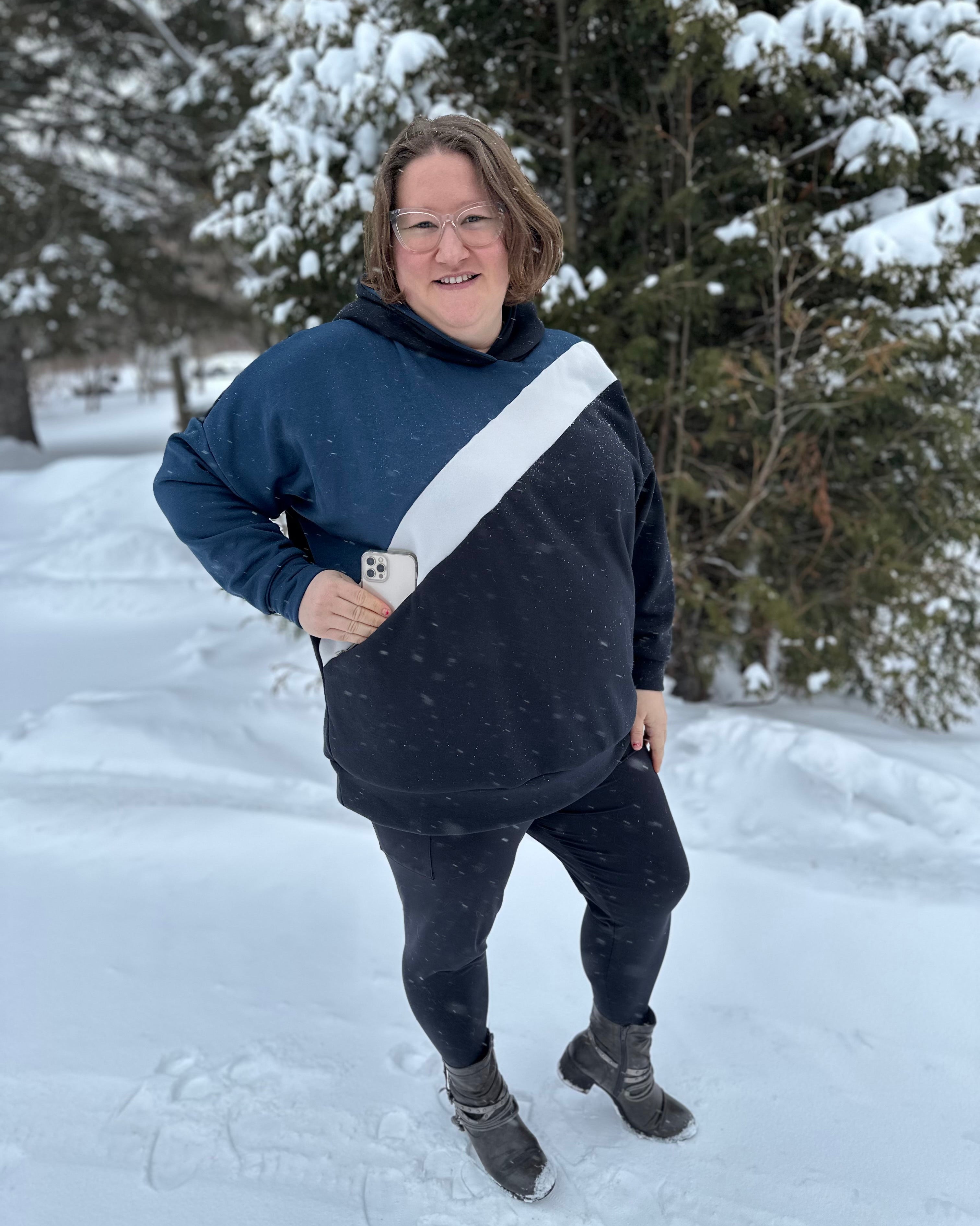 Person wearing a navy and gray hoodie with a white stripe, standing in a snowy landscape. Showing off the zip pocket with her cell phone going into it.