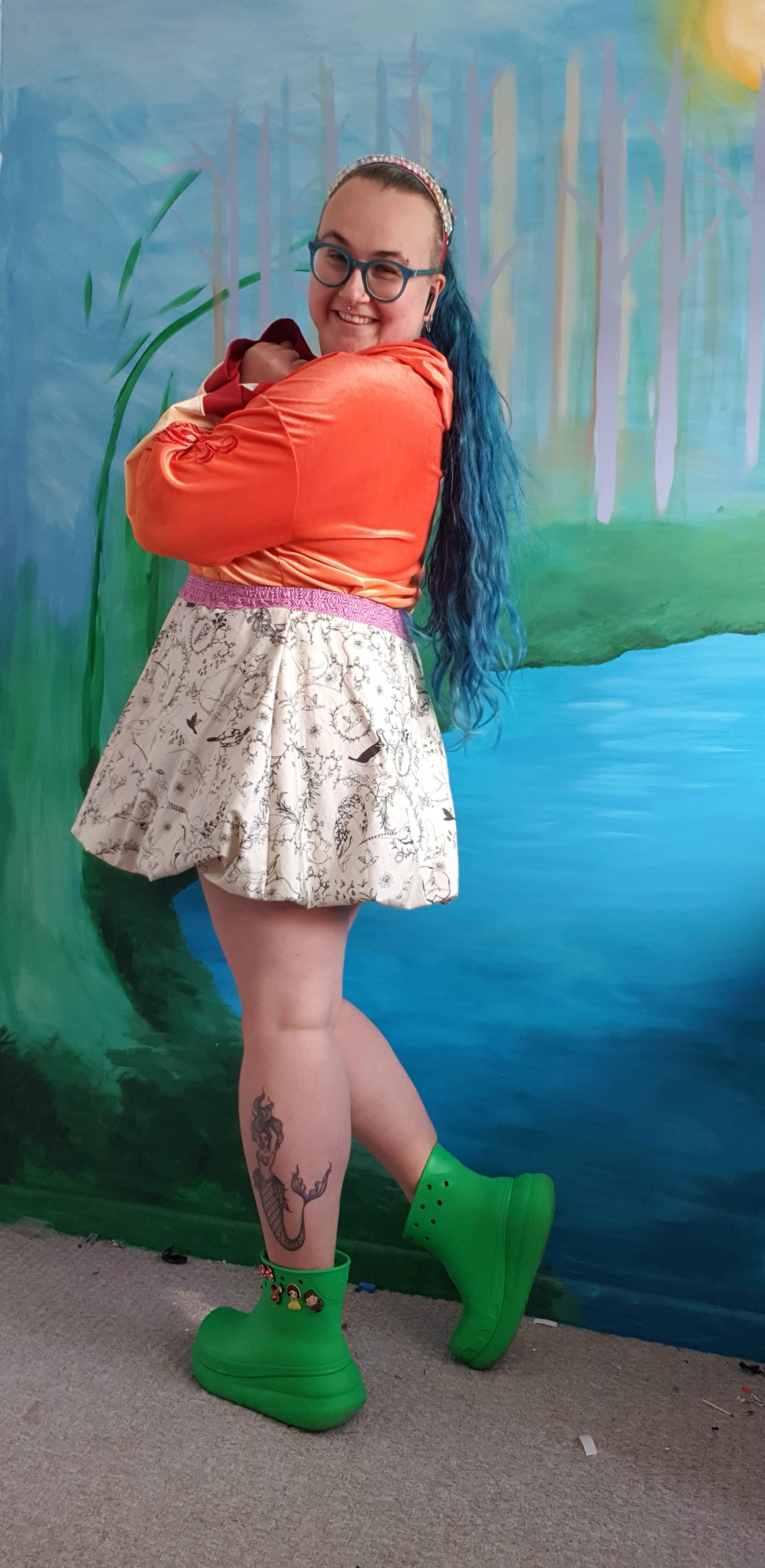 Person wearing an orange shirt, white mini bubble skirt, and green shoes against a colorful mural.