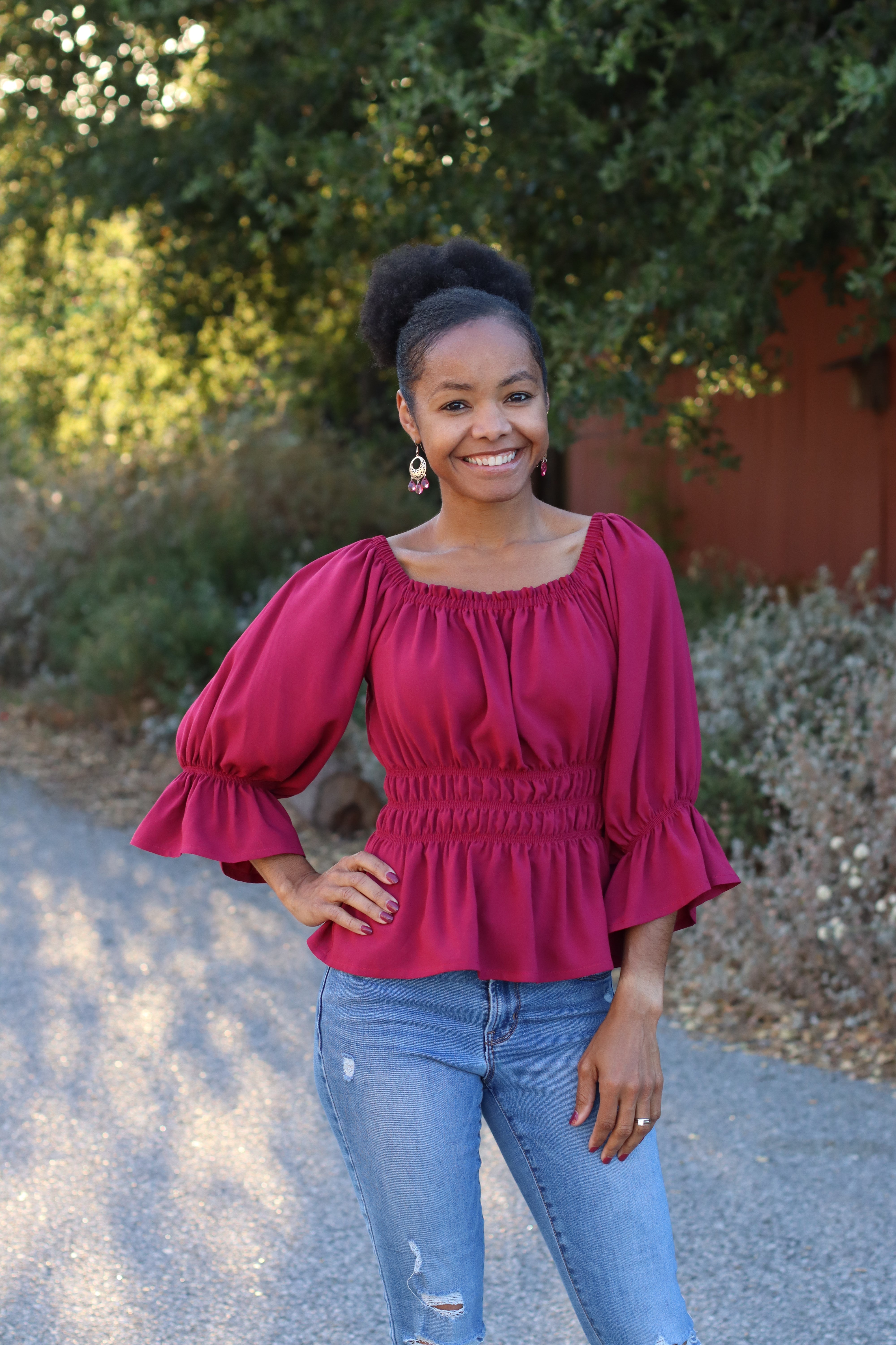 Woman wearing a red shirred blouse with 3/4 puff sleeves and blue jeans standing outdoors with greenery in the background. Made using the digital pdf sewing pattern by Ellie and Mac patterns.
