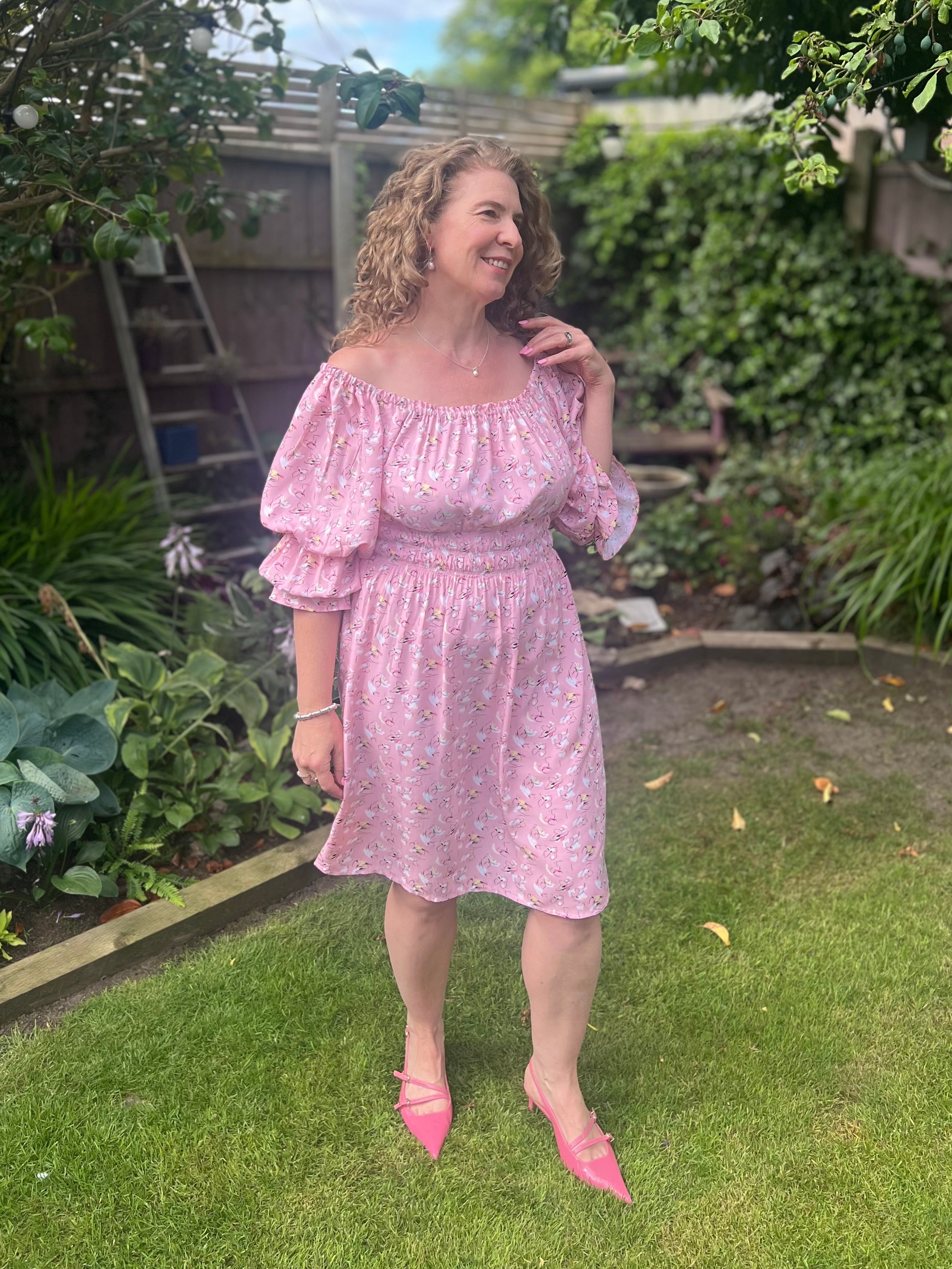 Woman in a pink knee-length shirred dress standing in a garden. The dress is worn off-shoulder with elbow length balloon sleeves. Sewing pattern by Ellie and Mac patterns.