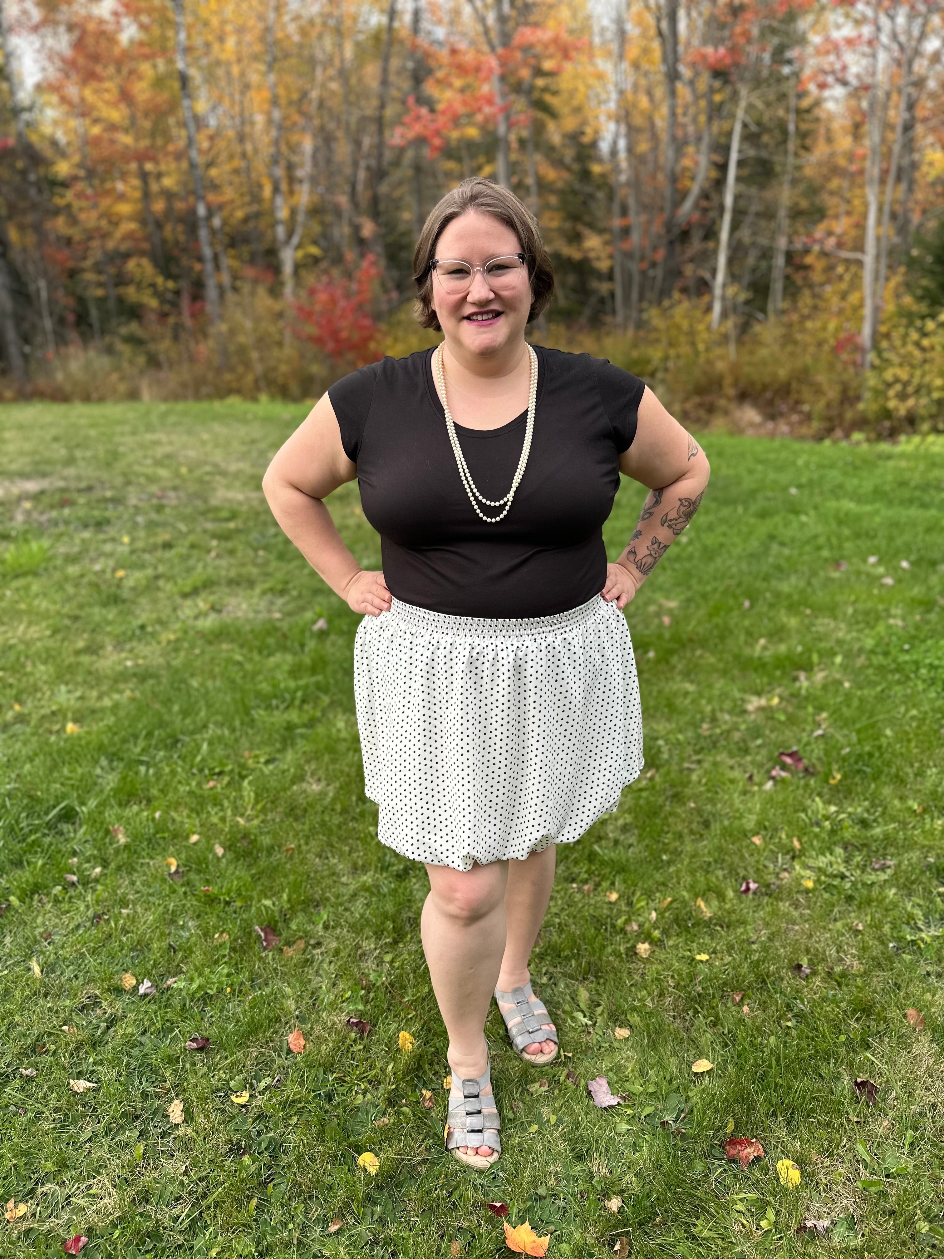 Person wearing a black top and white mini bubble skirt standing in a grassy area with trees in the background.
