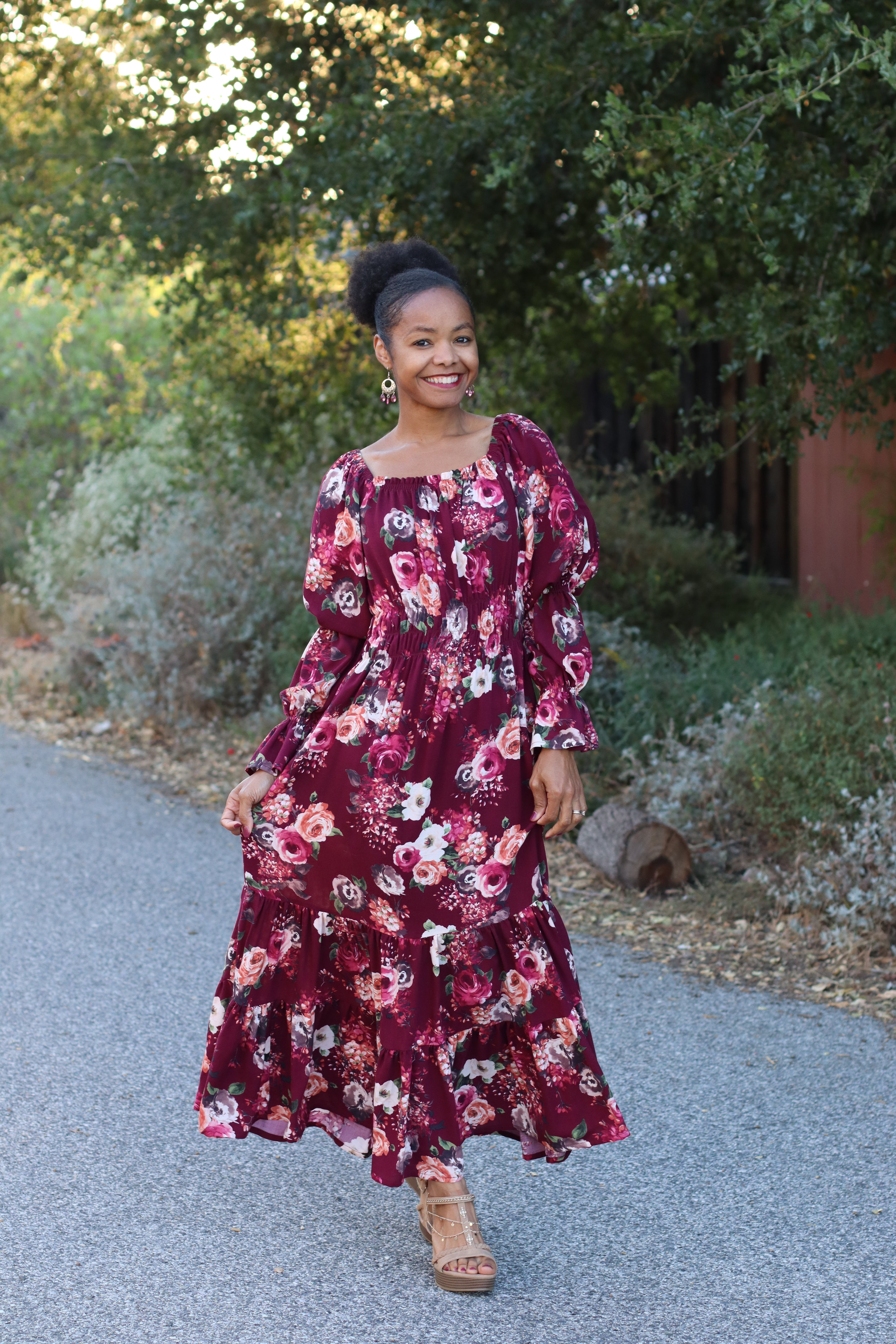 Woman in a floral midi length dress with long billowy statement sleeves, standing on a road with greenery in the background. The square peasant neckline screams cottage core style. Inclusive sizing sewing pattern.