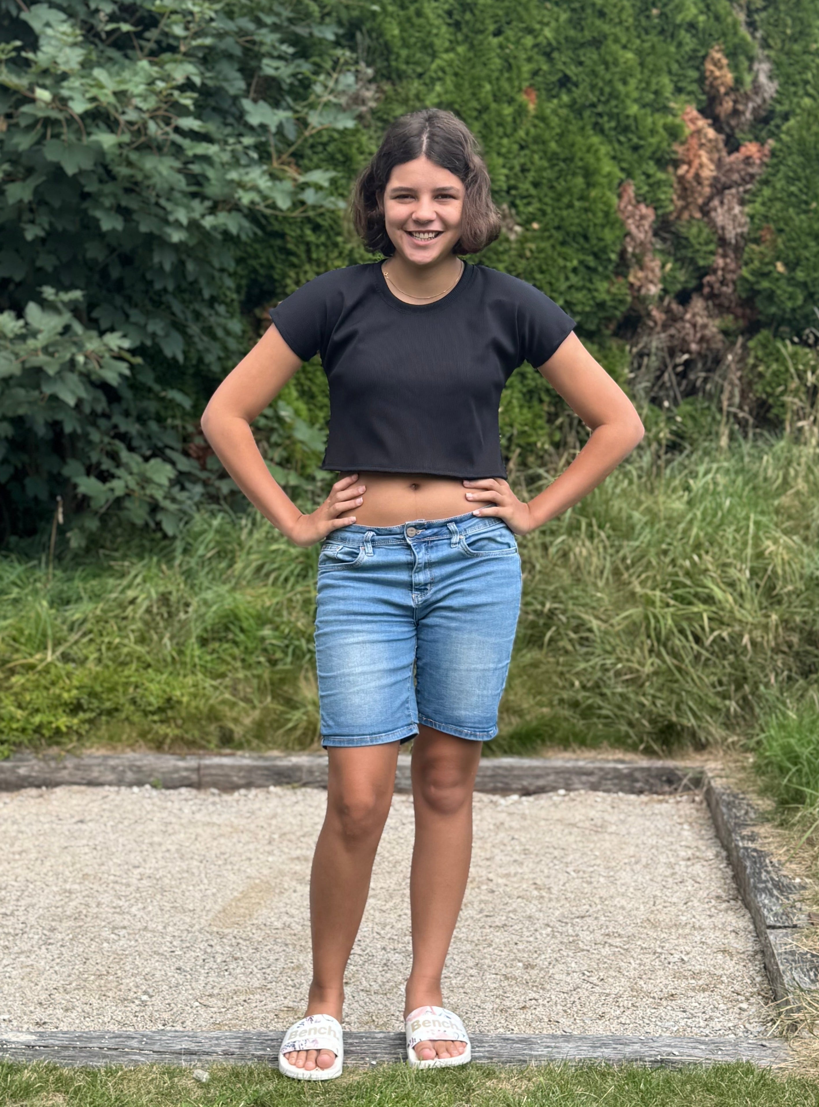 Person wearing a black crop top and denim shorts standing outdoors with greenery in the background. Top made using the Kayla Crop Top PDF sewing pattern.