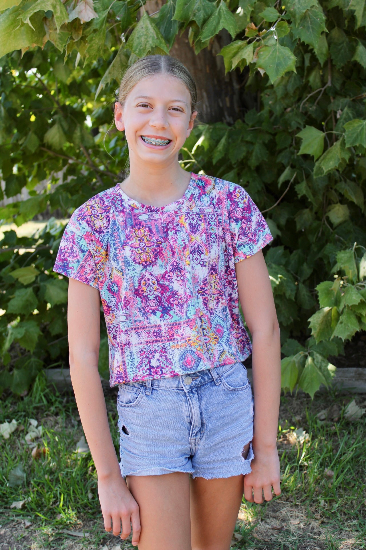 Teen girl wearing a colorful floral top and denim shorts standing in front of green foliage.
