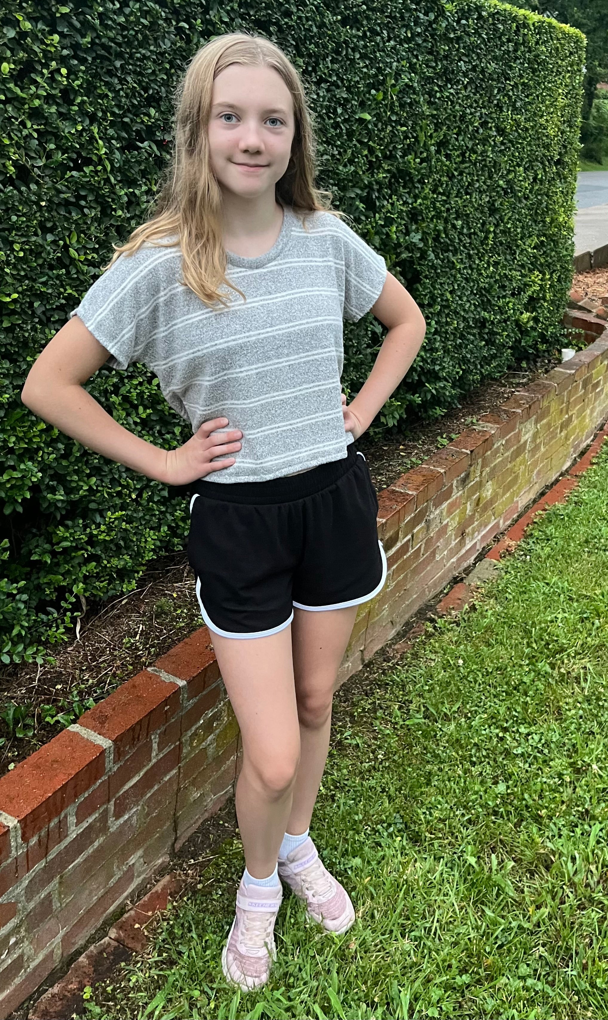 Young girl standing outdoors on a grassy area with a hedge in the background