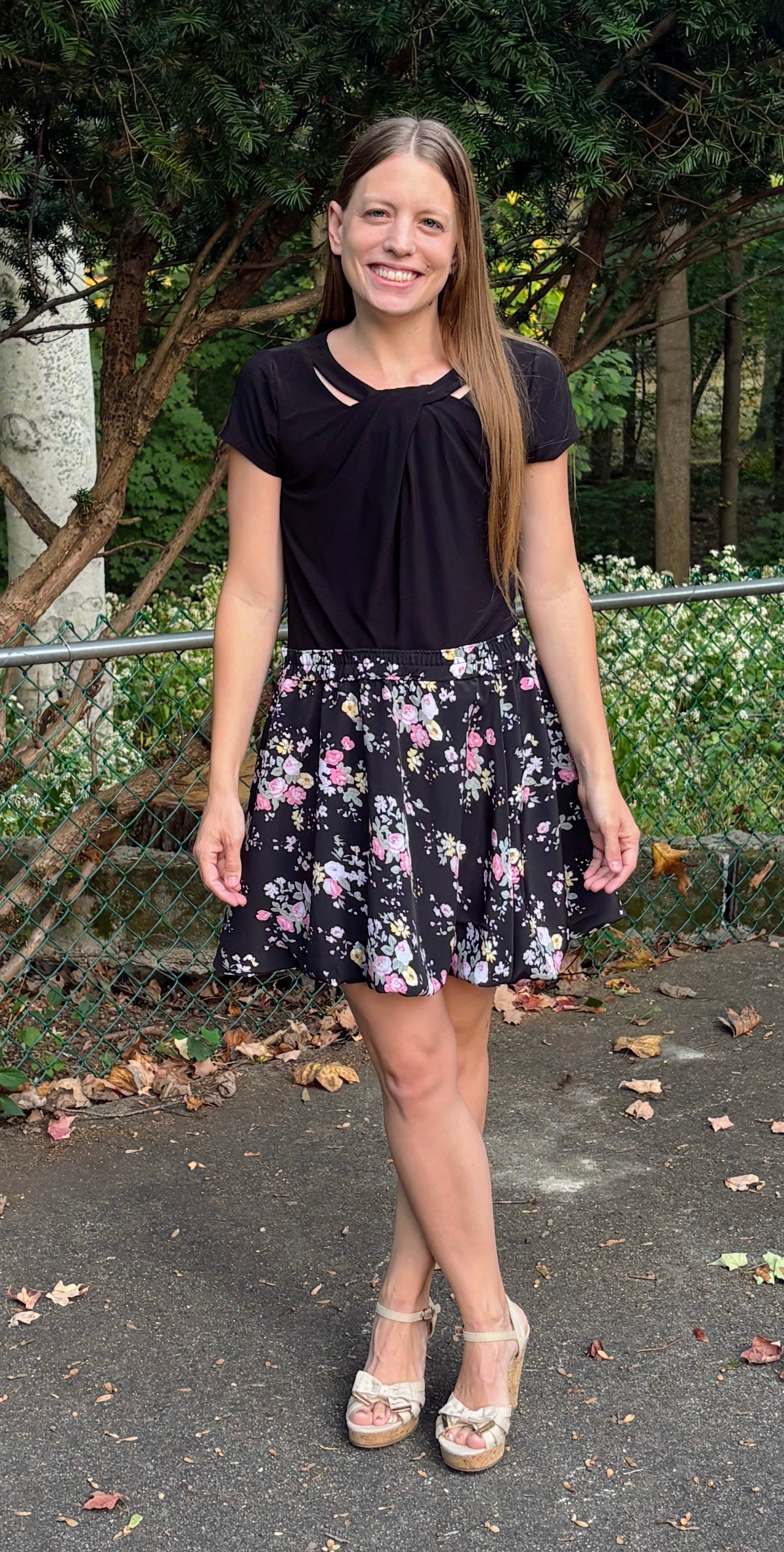 Woman wearing a black top and floral bubble skirt standing outdoors with trees and bushes in the background.
