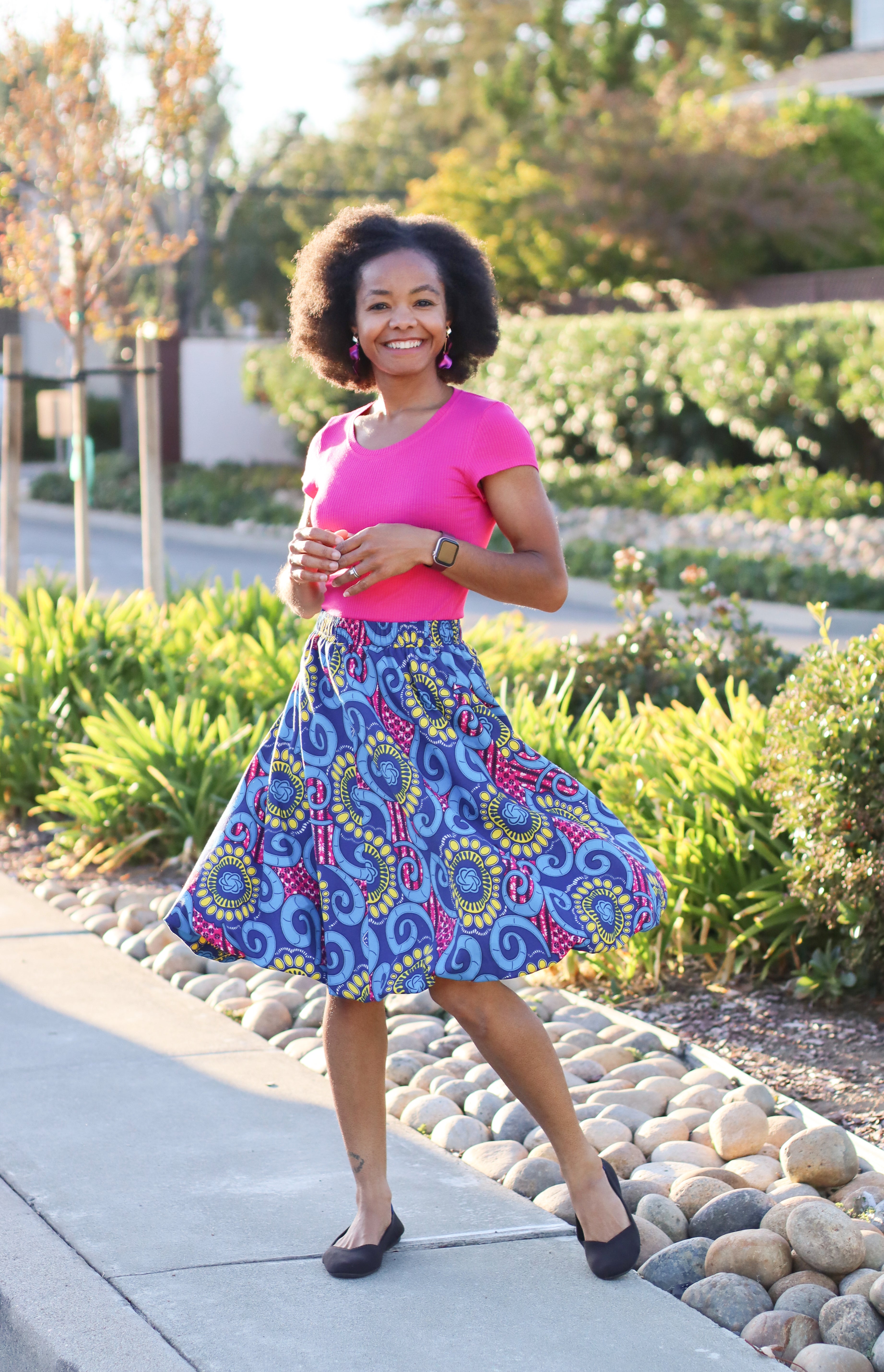 Woman in a bright pink top and colorful skirt standing outdoors on a sunny day. The bubble skirt is swishing back and forth. The Model is smiling and loving her skirt.