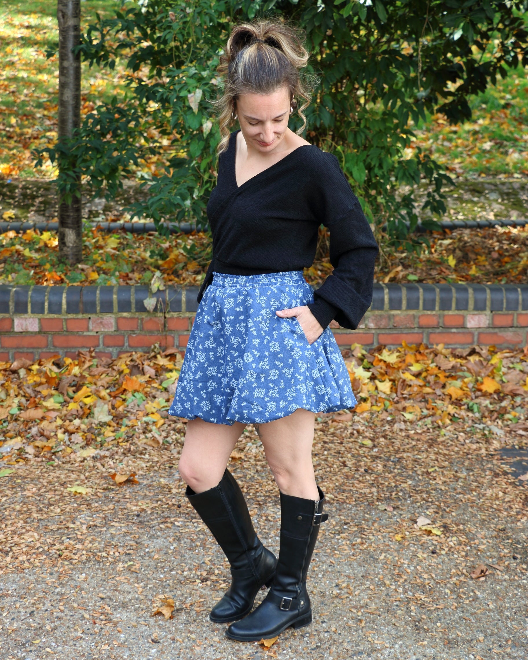 Woman wearing a black bishop sleeve top, blue mini bubble skirt, and black boots standing outdoors with fallen leaves on the ground.