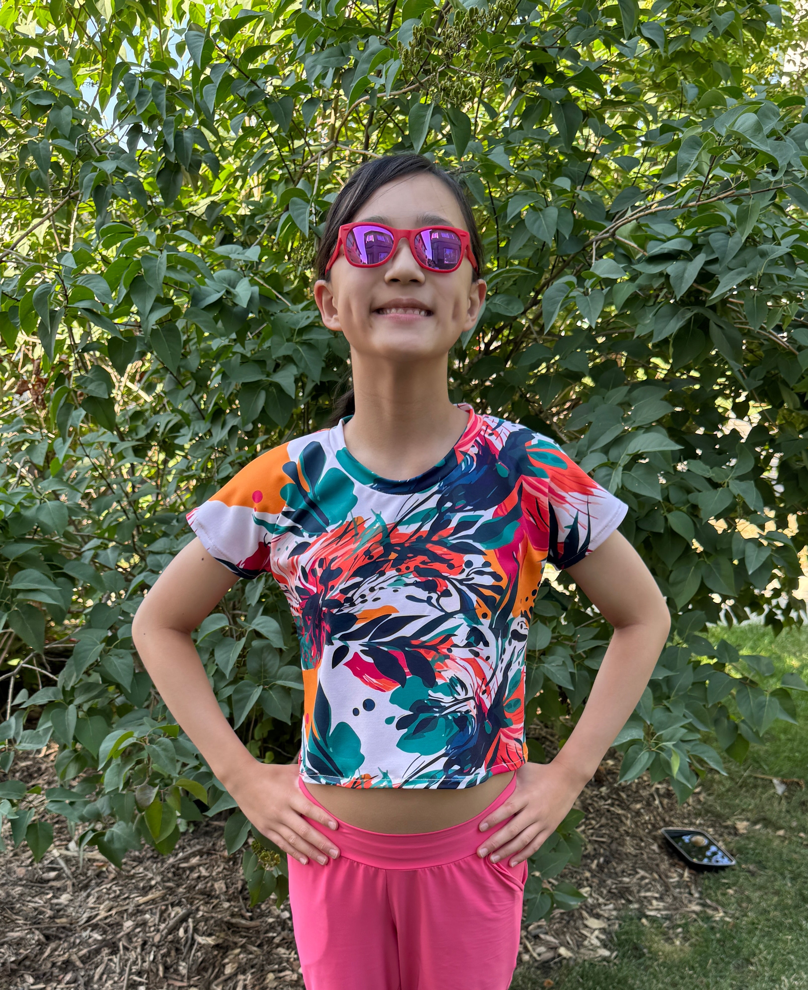 Person wearing a colorful floral top and pink shorts standing outdoors with greenery in the background