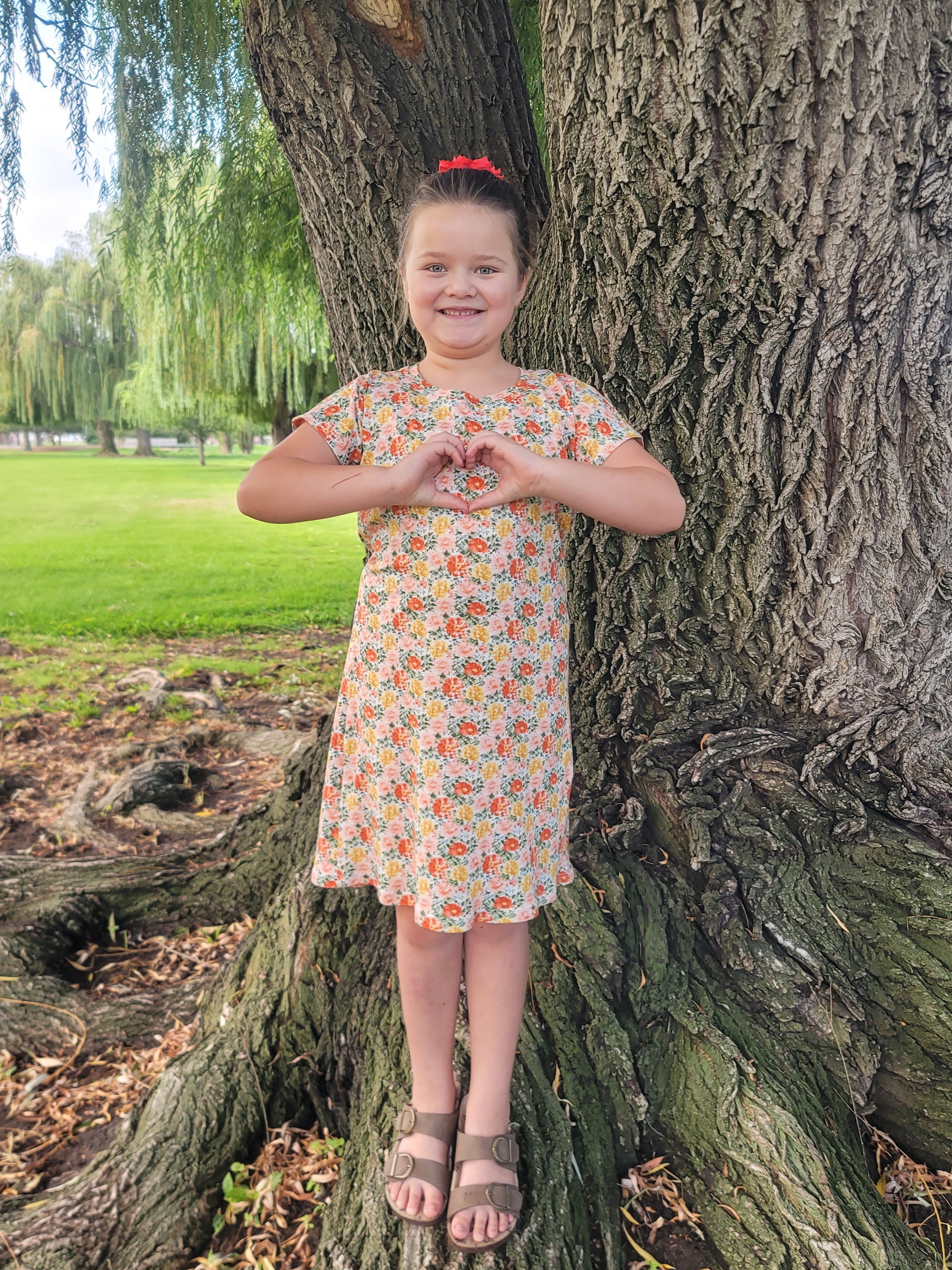 Person in a floral dress standing between two trees with a grassy area and trees in the background.