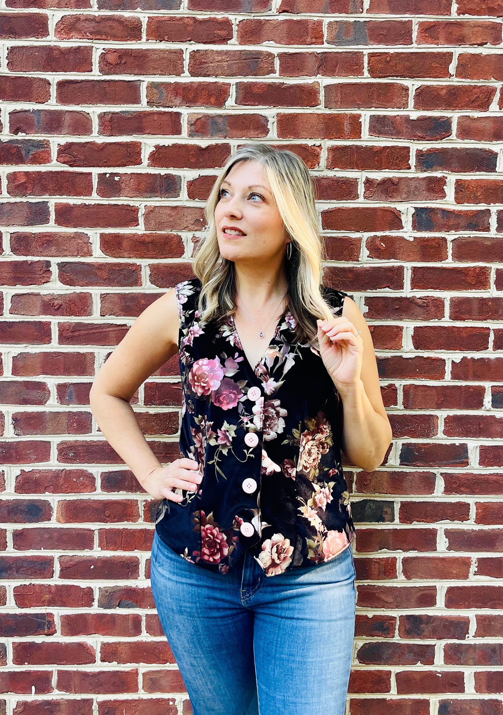 Woman wearing a floral on black velvet vest with pink buttons and jeans. Standing by a brick wall. PDF sewing pattern for beginners.