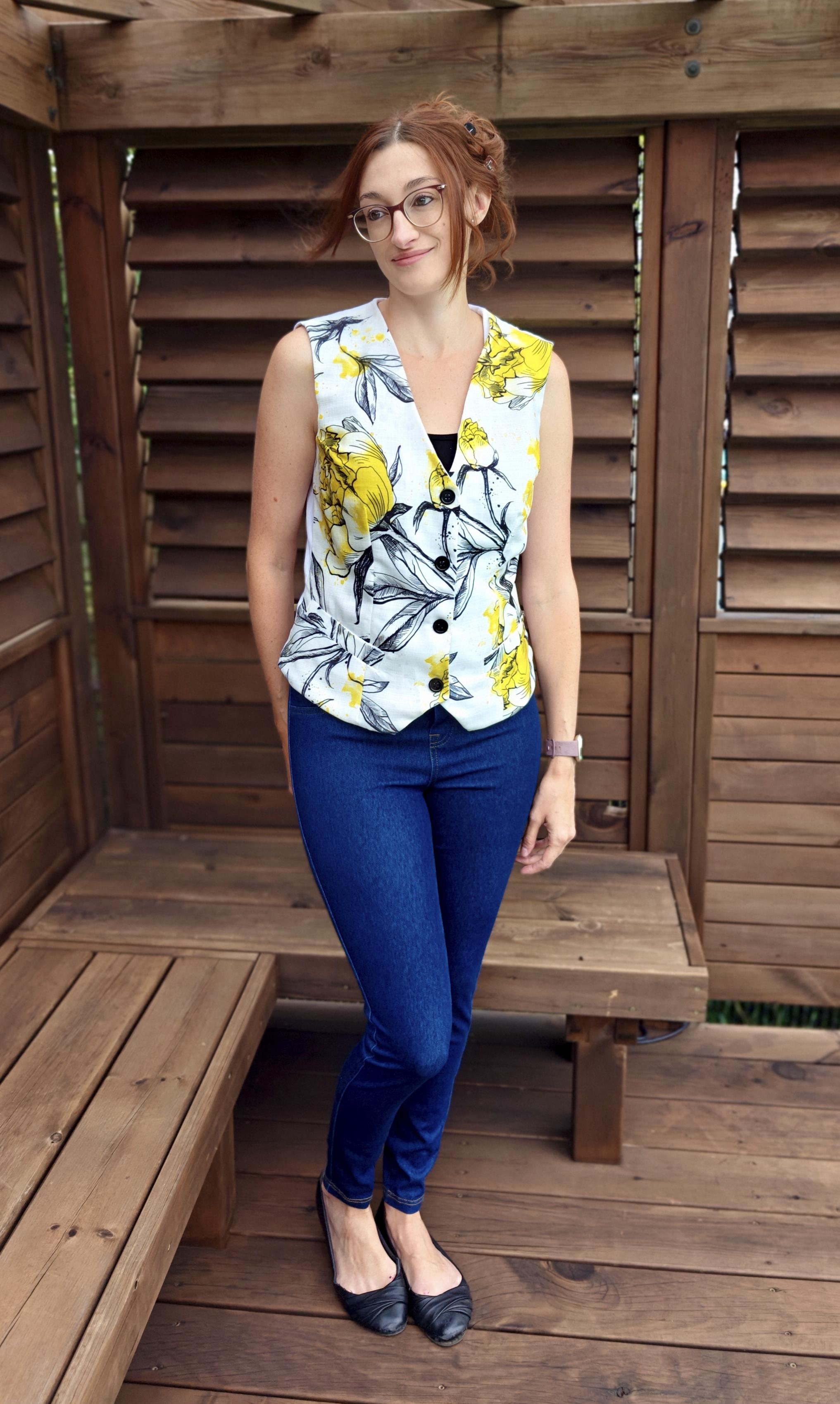 Woman wearing a floral vest with welt pockets and denim skinny jeans standing on a wooden deck.