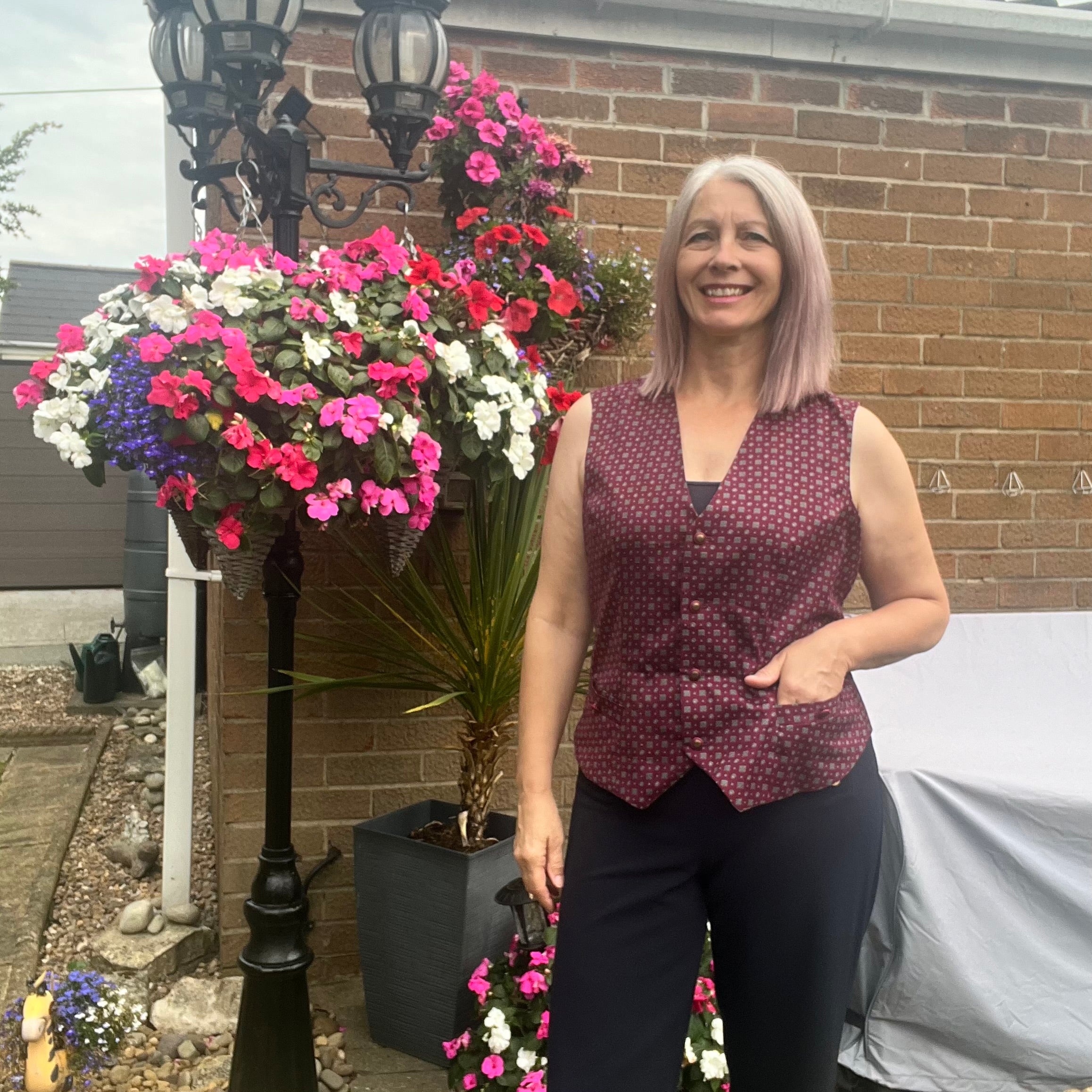 Woman wearing a burgundy vest and black pants standing by a brick wall and flowers outside. The vest is the high hip length option made from the Veronik Waistcoat Vest sewing pattern by Ellie and Mac Sewing Patterns.