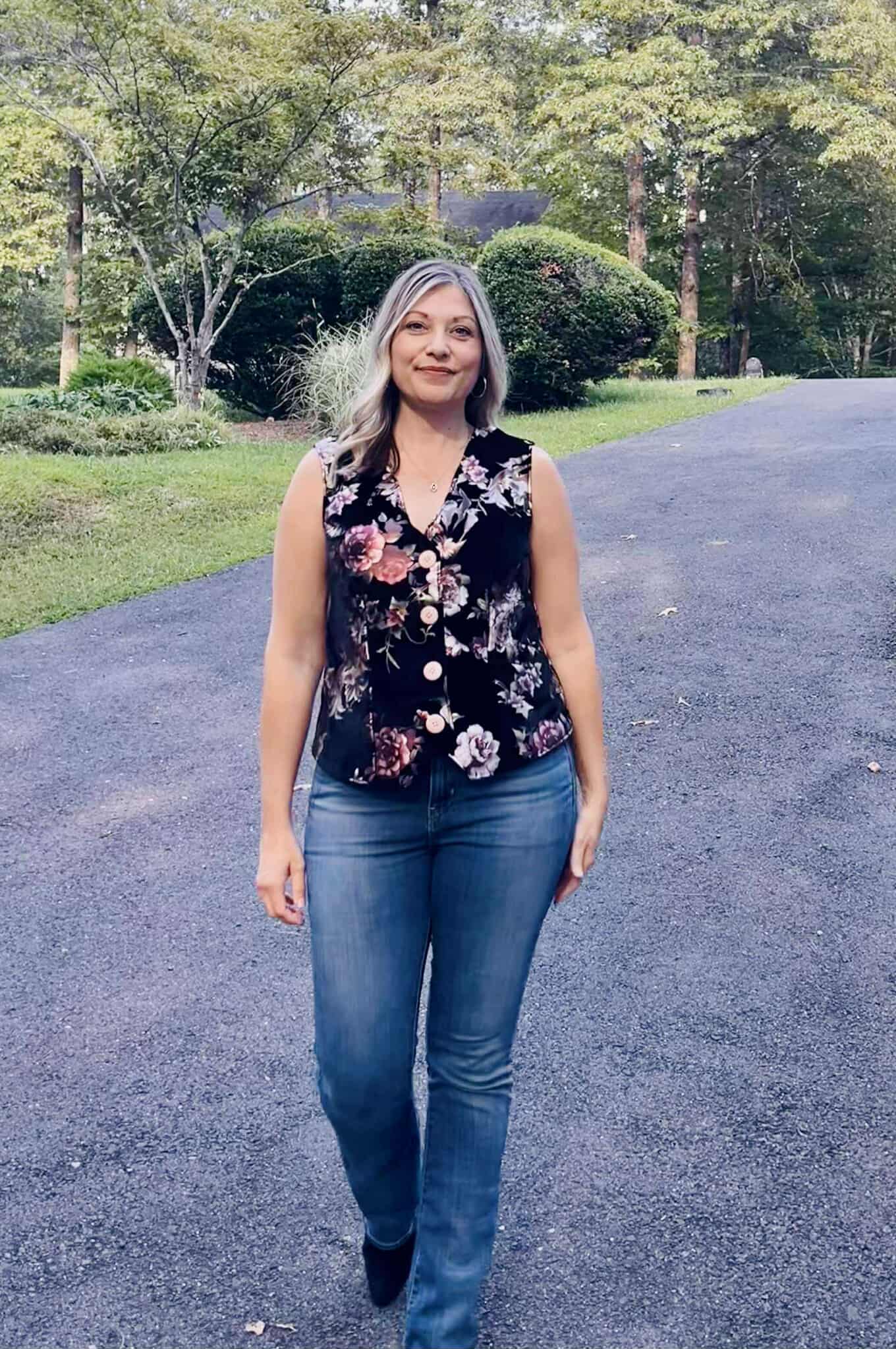 Woman walking outside in a paved driveway wearing jeans, boots, and a black velvet vest with floral print. The vest is high hip length. Made using the Ellie and Mac Veronik sewing pattern.