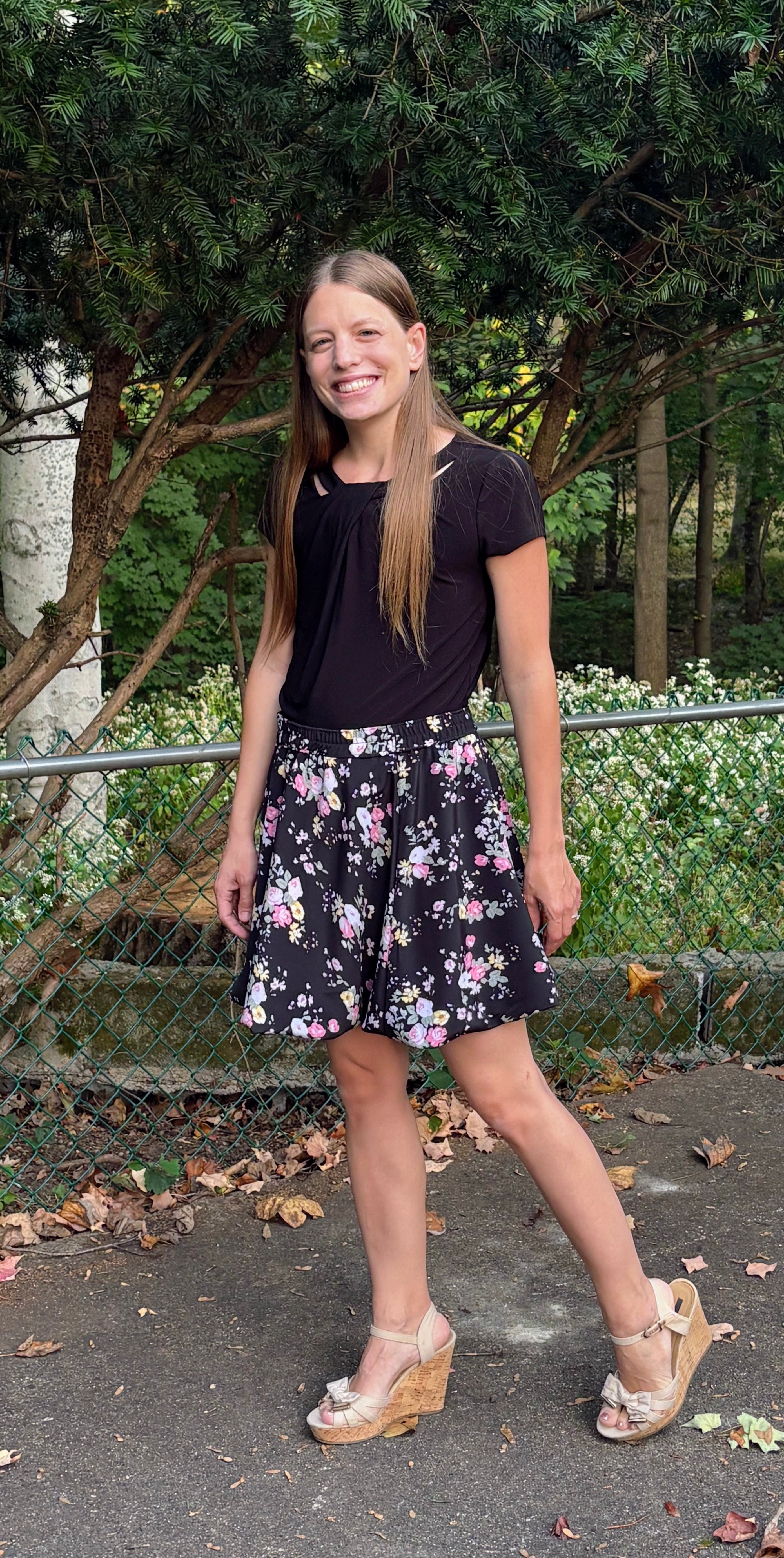 Woman wearing a black top and floral bubble skirt standing outdoors with greenery in the background.