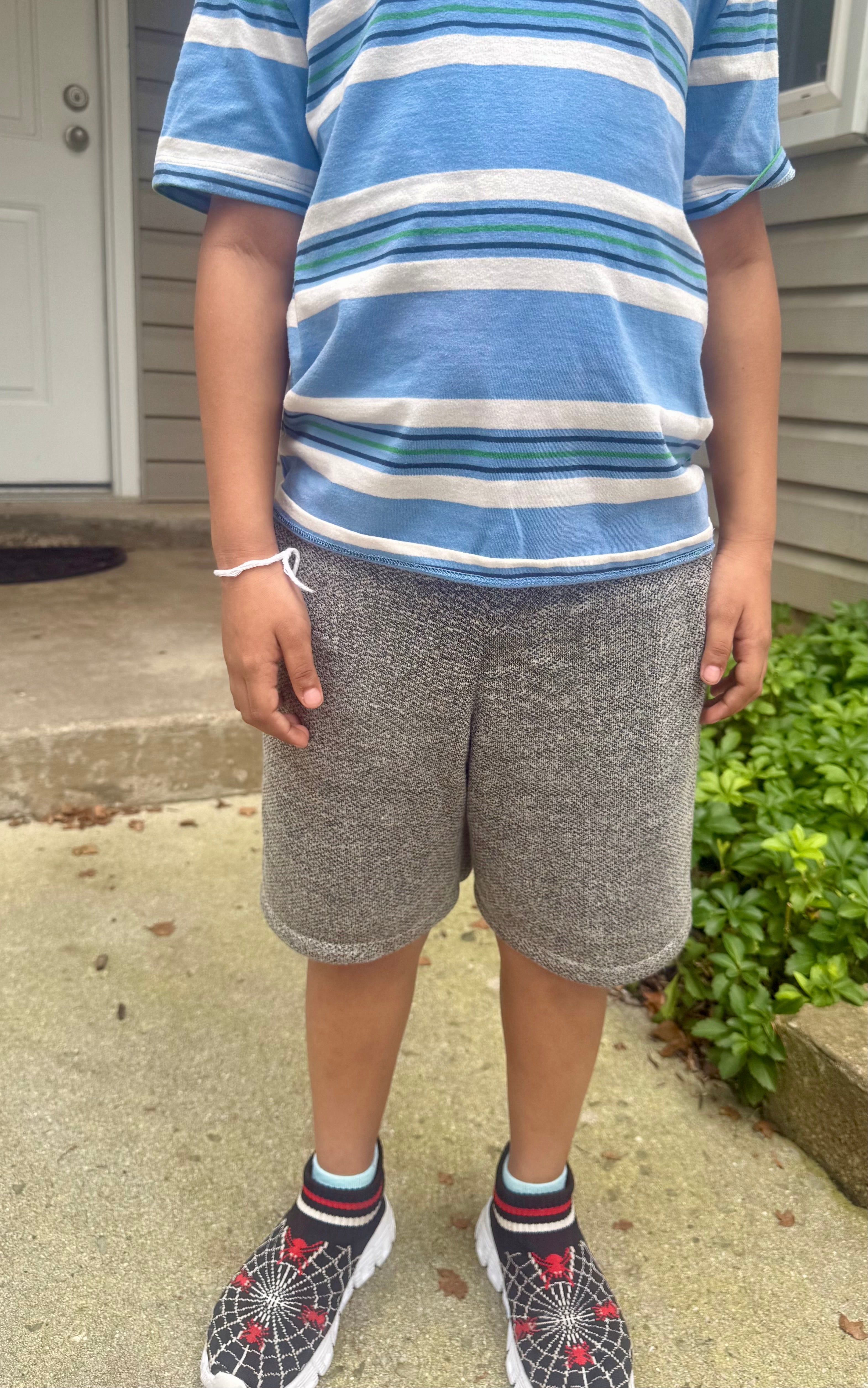 Person wearing a blue and white striped shirt and gray shorts standing on a sidewalk. Shorts are Above knee length.