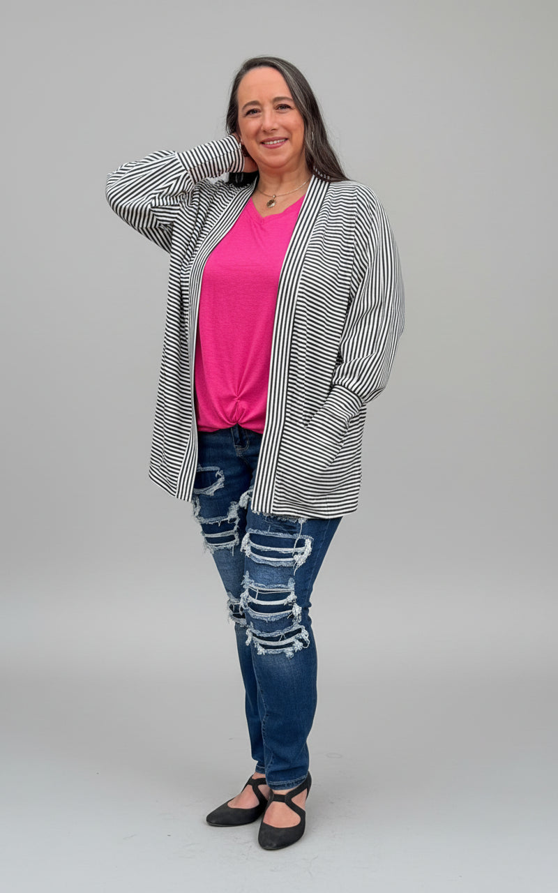 Woman wearing a striped cardigan, pink shirt, and ripped jeans standing against a green bush. PDF sewing pattern.