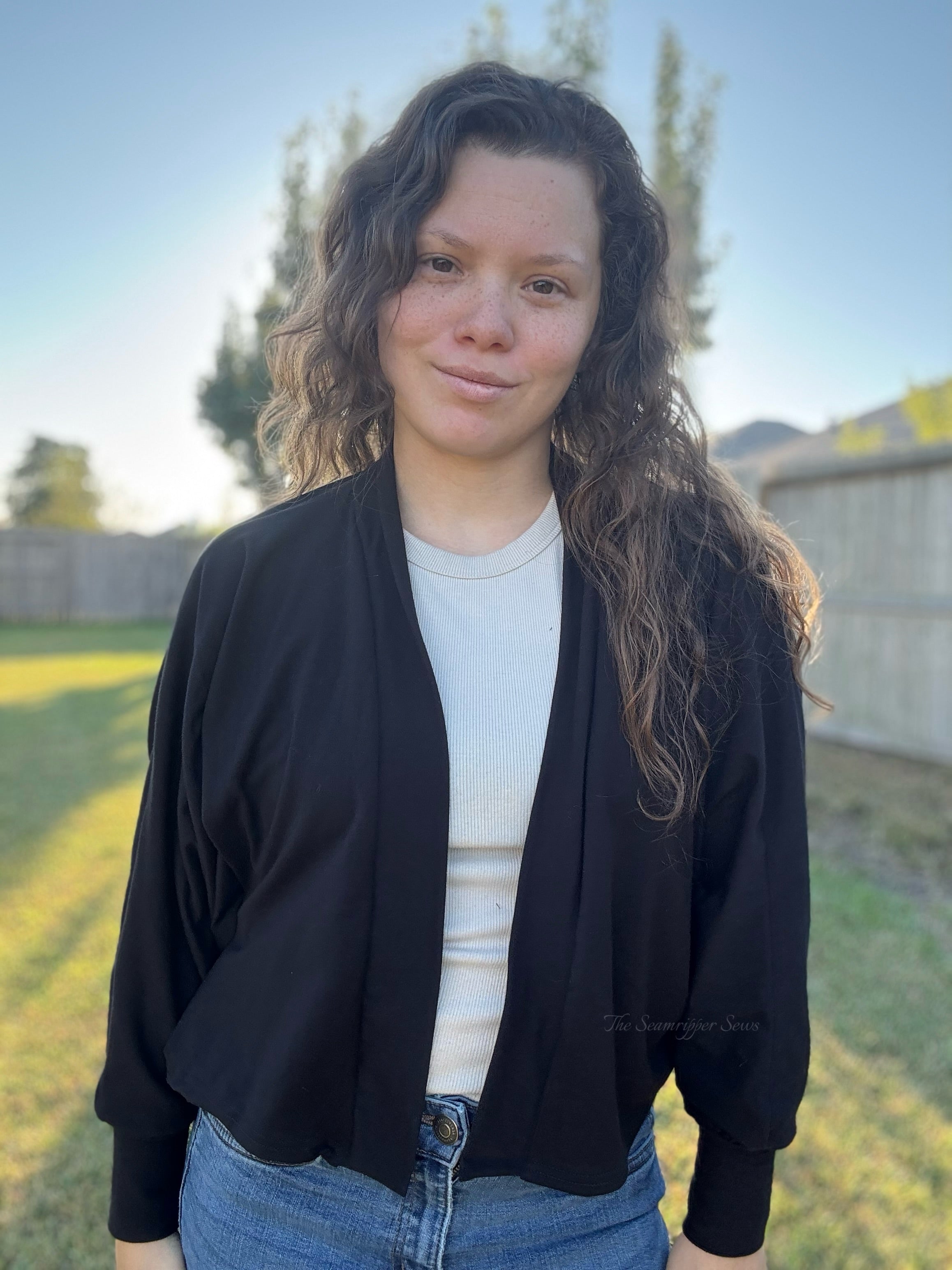 Person wearing a black cropped batwing cardigan jacket over a white shirt with a blurred outdoor background