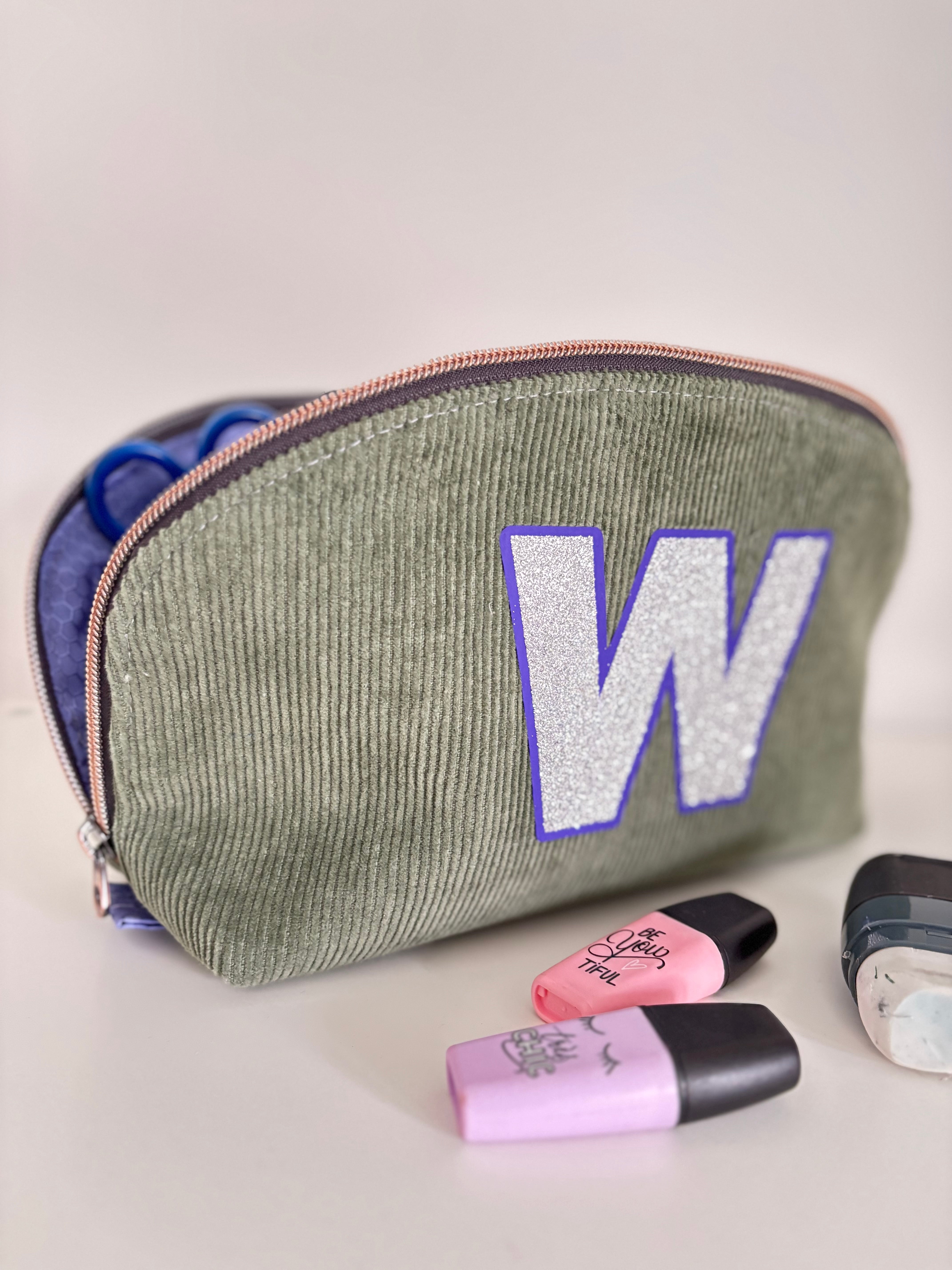 Green cosmetic bag with a purple 'W' and makeup items on a light gray background made from Ellie and Mac sewing pattens.