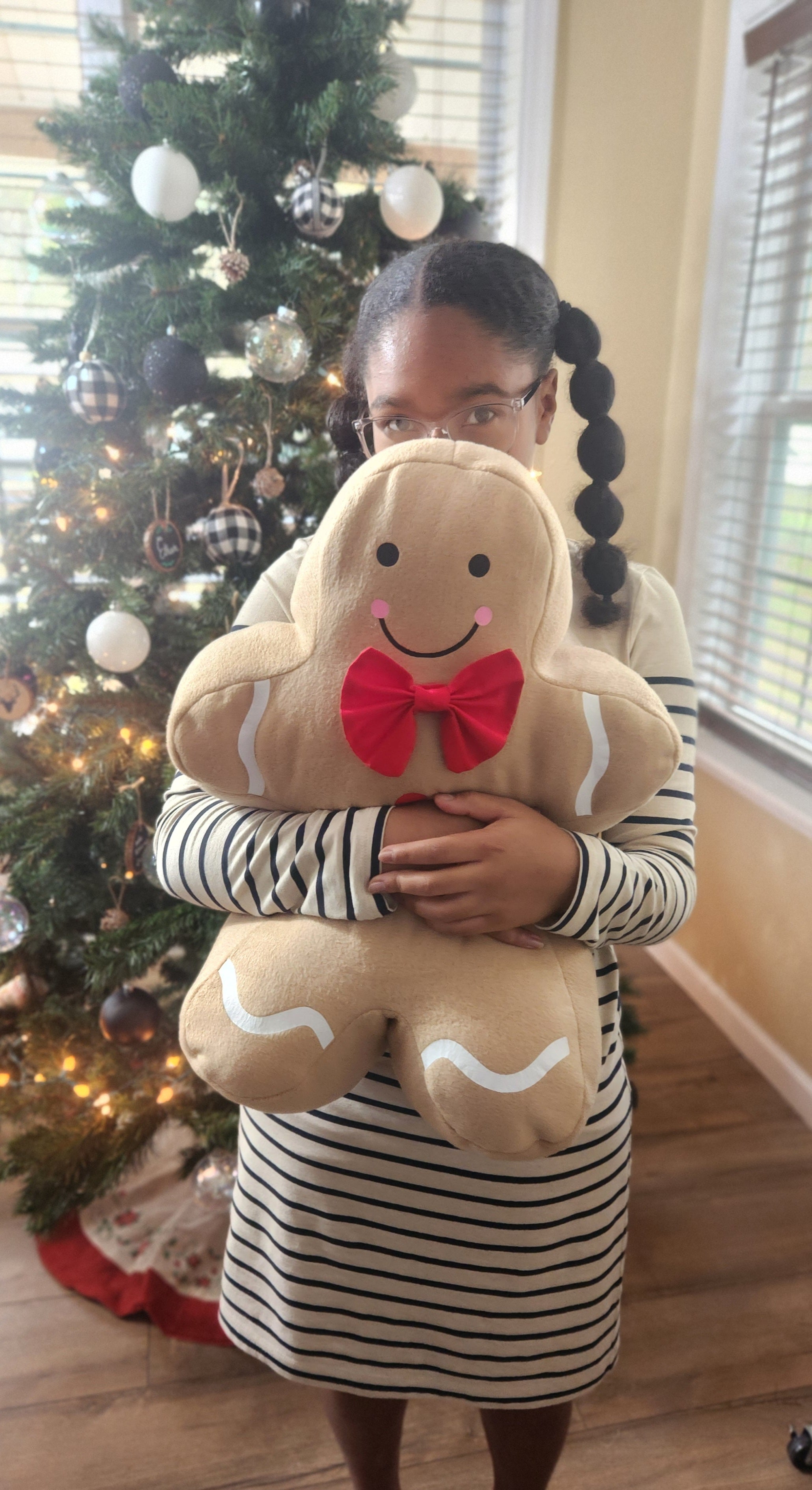 Jolly Gingerbread pillows displayed in a child’s arms in front of a Christmas Tree with holiday décor.