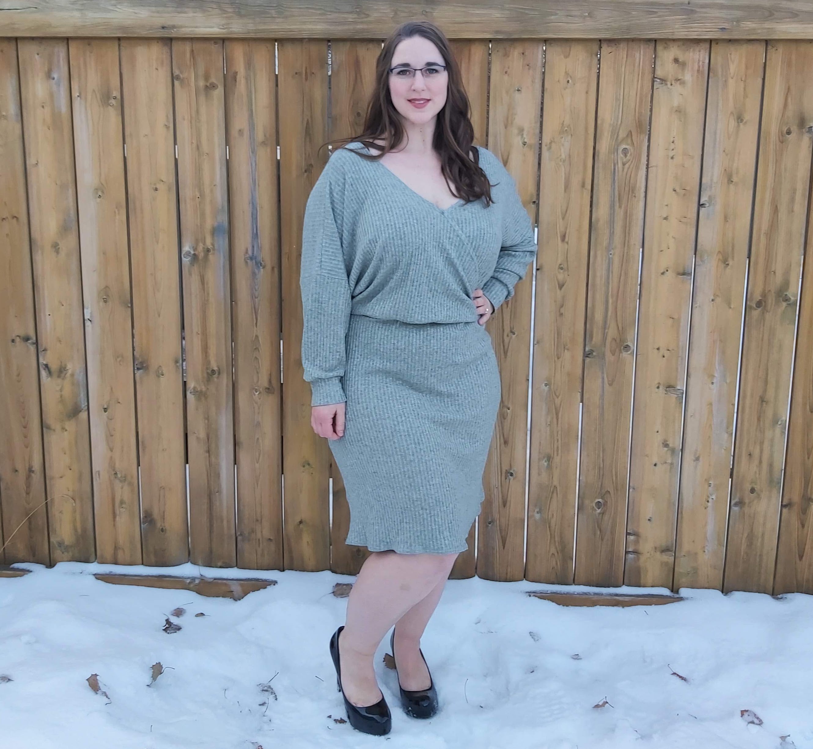 Woman wearing a grey crossover dolman sweater dress standing in front of a wooden fence with snow on the ground.