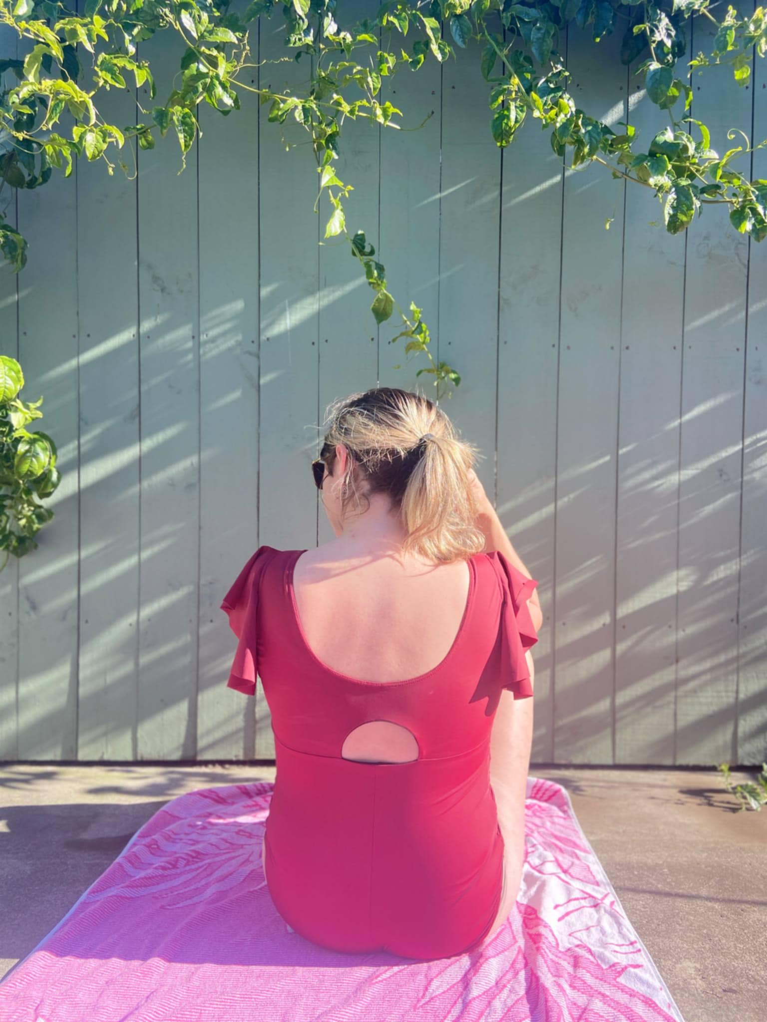Person wearing a red swimsuit with cut-out back sitting on a pink blanket against a white fence.