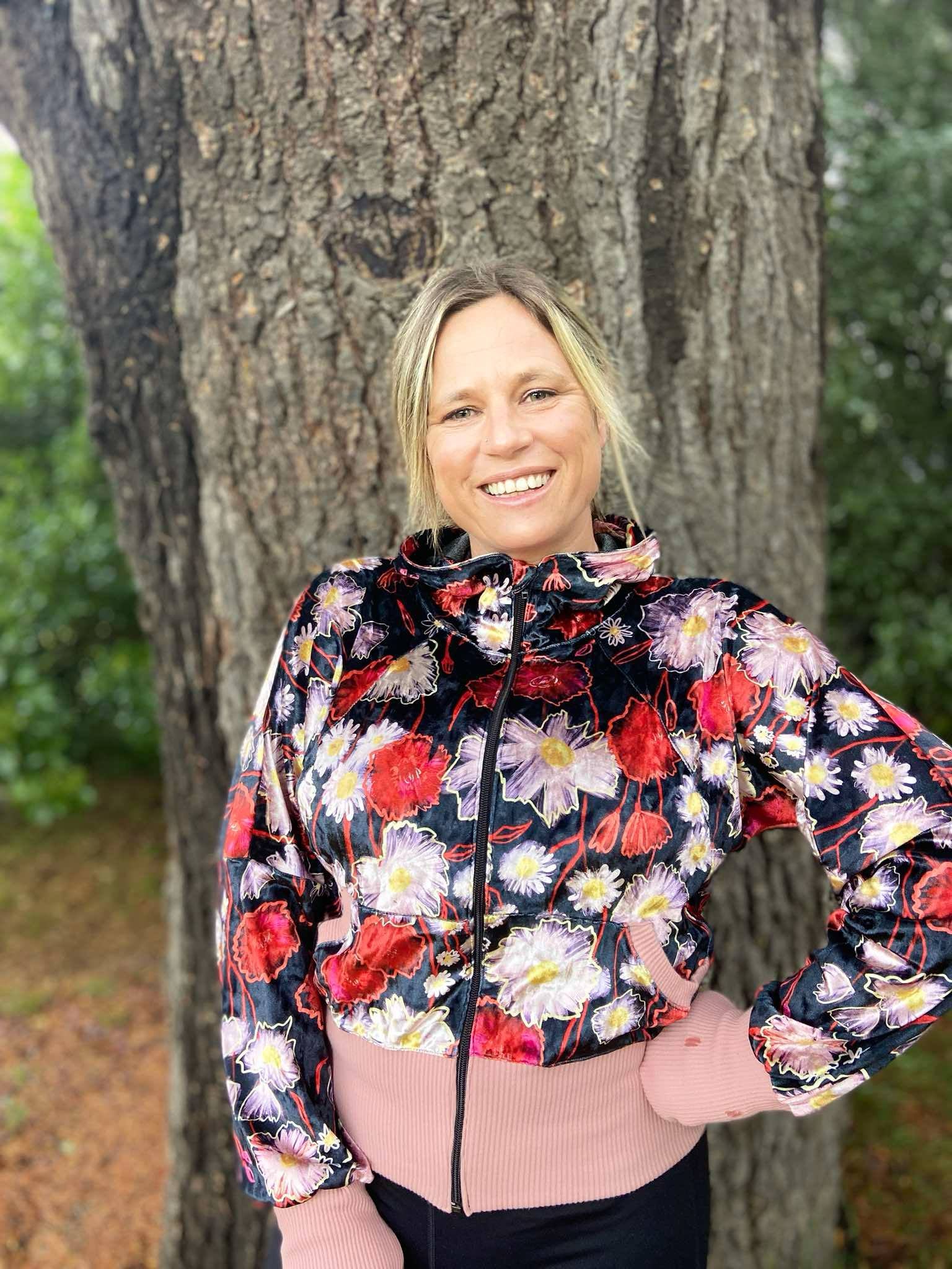 Woman outside modeling a bright floral athleisure hooded jacket with zip closure, kangaroo pocket detail, and thumbhole cuffs. Easy pdf sewing pattern.