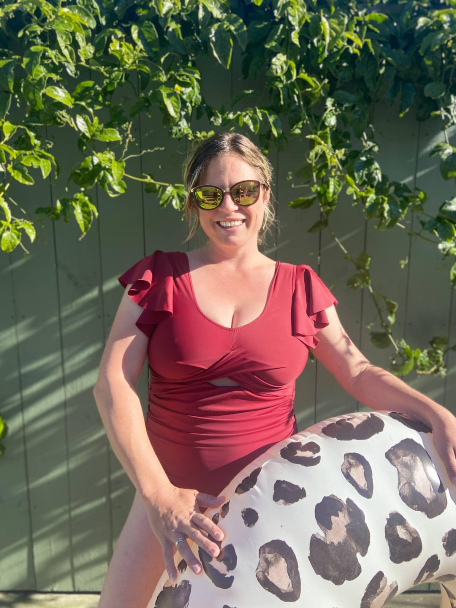 Woman in a red swimsuit holding a leopard print pool float against a green wall with foliage. The swimsuit was made with the tropical twist swimsuit pdf sewing pattern. It features cut outs in front and back.