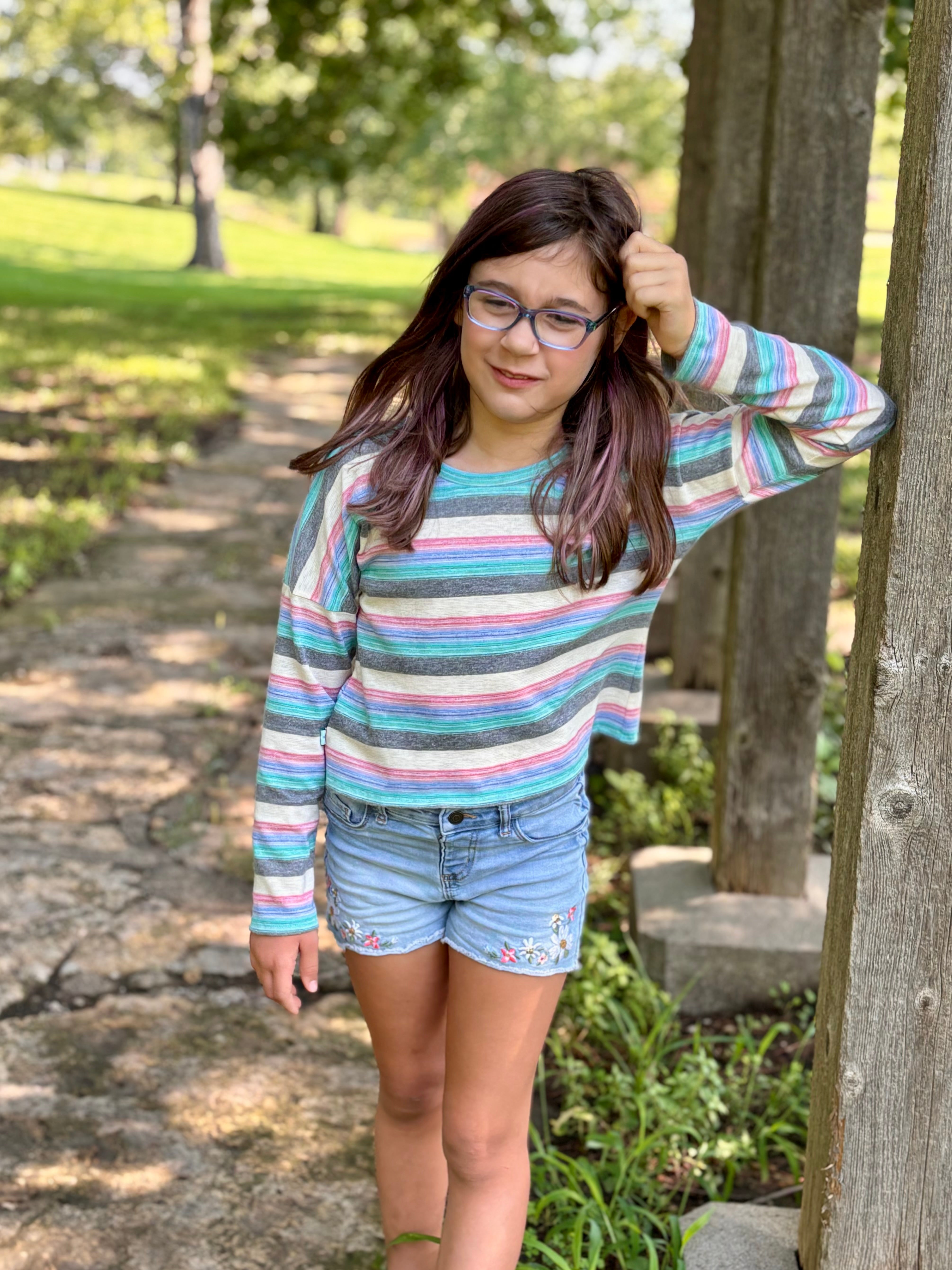 Young girl in a striped dolman shirt and denim shorts standing outdoors near a tree. Top made using Ellie and Mac's Kayla Crop Top Pattern.