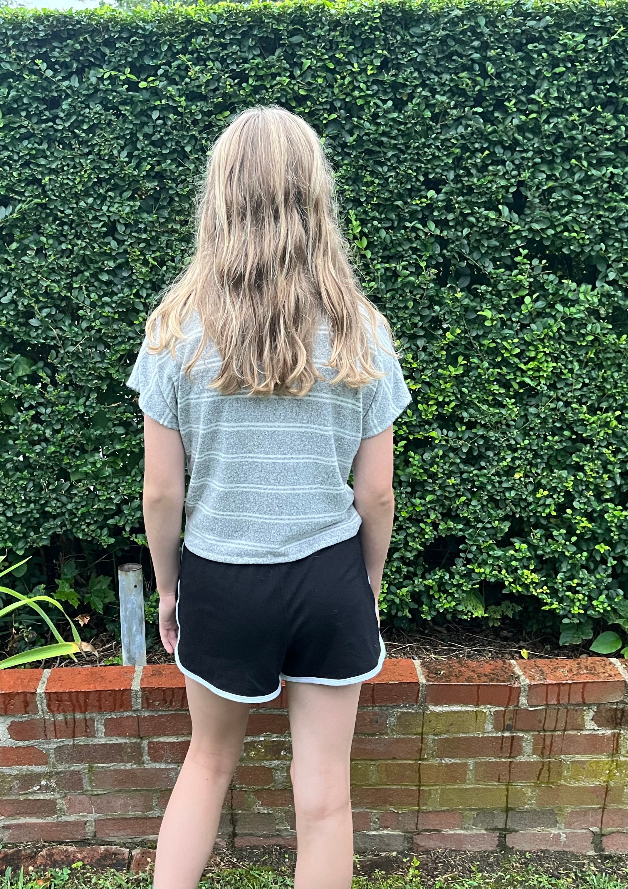 Person standing in front of a hedge with a brick wall and plants in the foreground